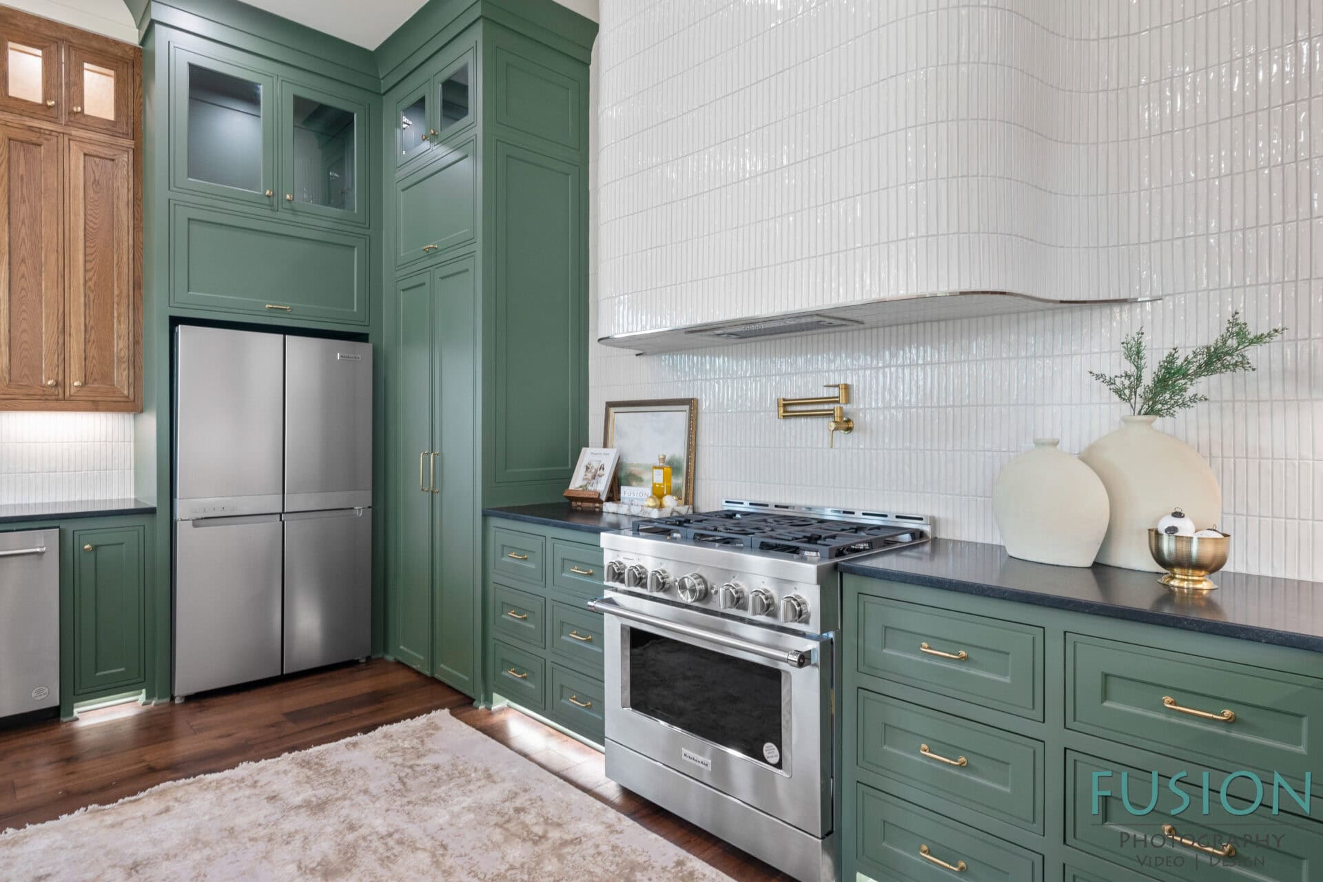 A modern kitchen with green cabinets, stainless steel refrigerator, and gas stove. White textured tile backsplash, black countertops, and decorative vases are visible. A framed photo and small plant are on the counter.