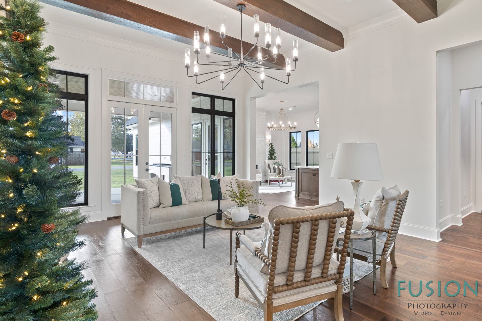 A bright living room with white walls, wood ceiling beams, and a large window. Furnishings include a gray sofa, two wooden chairs, a rug, a coffee table, and a decorated Christmas tree. Fusion Photography logo is visible.