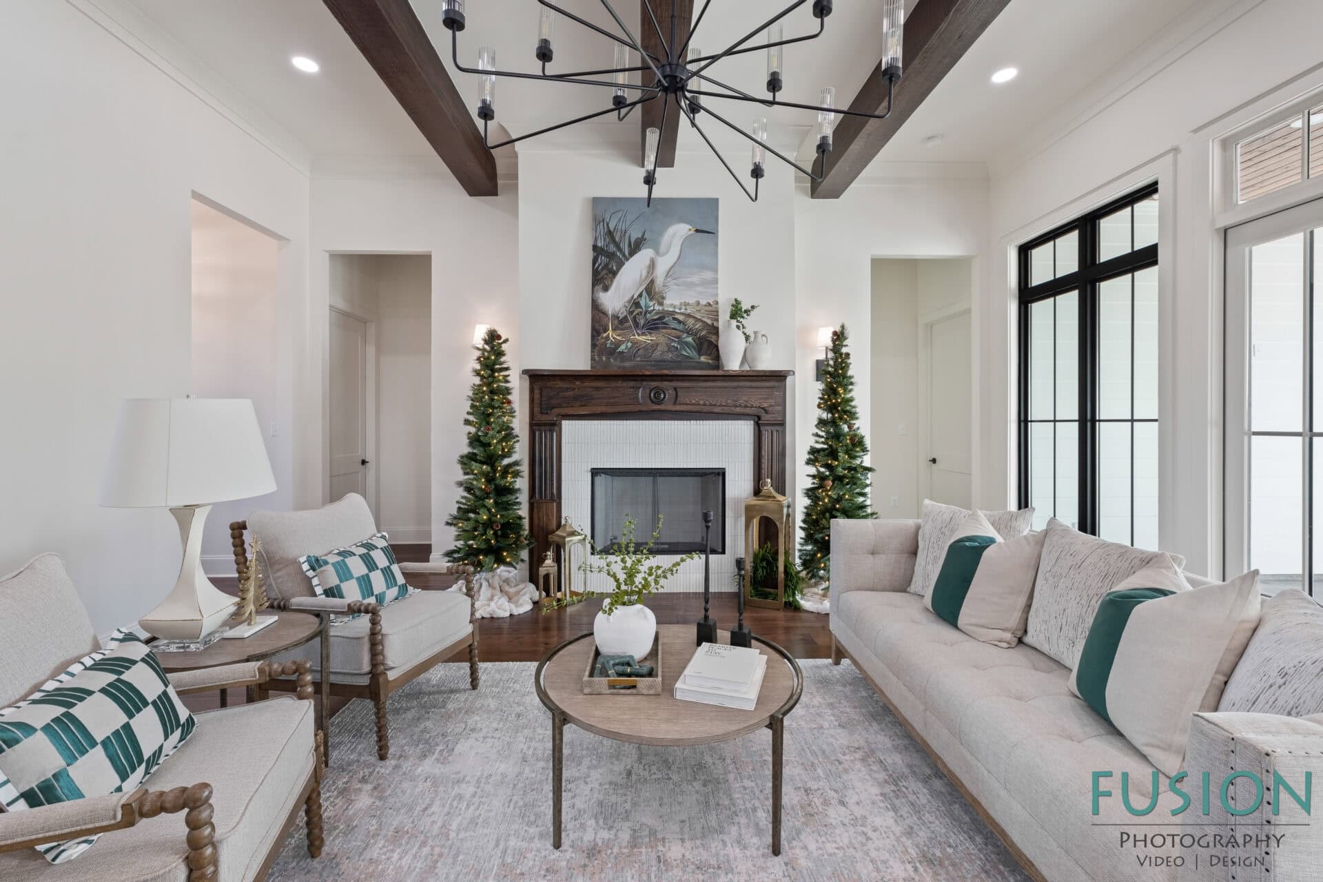 A bright living room with a white sofa, striped accent chairs, a wooden coffee table, a fireplace with a painting above it, two decorated trees, and large windows letting in natural light.