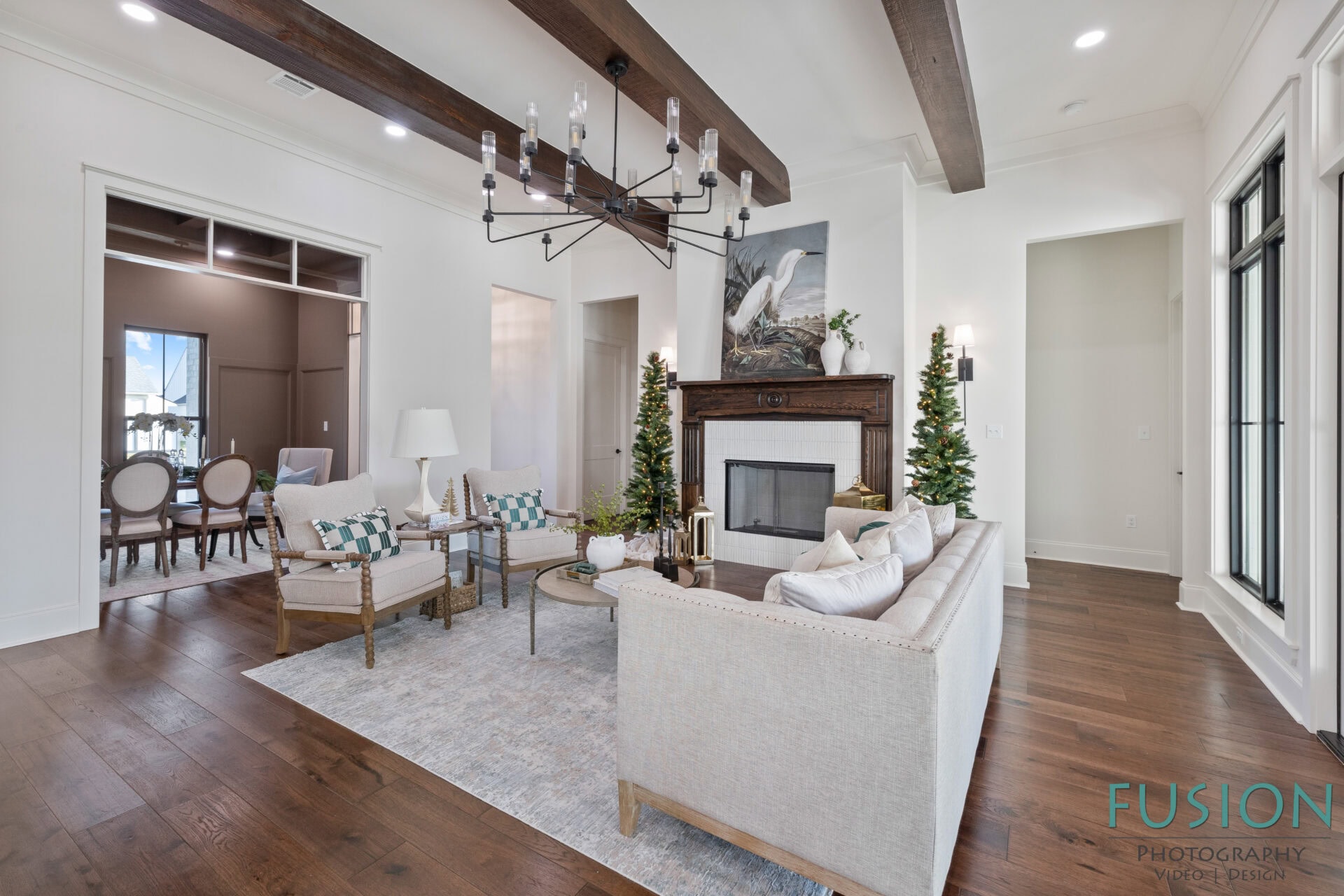 A modern living room with exposed wooden beams, white walls, and wooden floors. There is a fireplace with two small trees on each side, light-colored sofas, a coffee table, and a chandelier overhead.