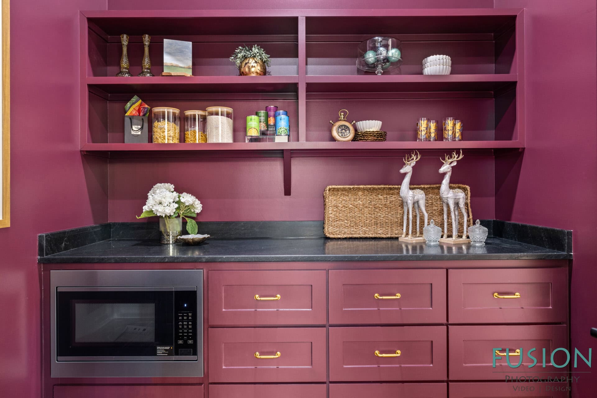 A burgundy kitchen wall with open shelves displaying jars, cans, and decor; a black countertop holds a microwave, a basket, two decorative reindeer, and a vase with white flowers. There are drawers with gold handles.