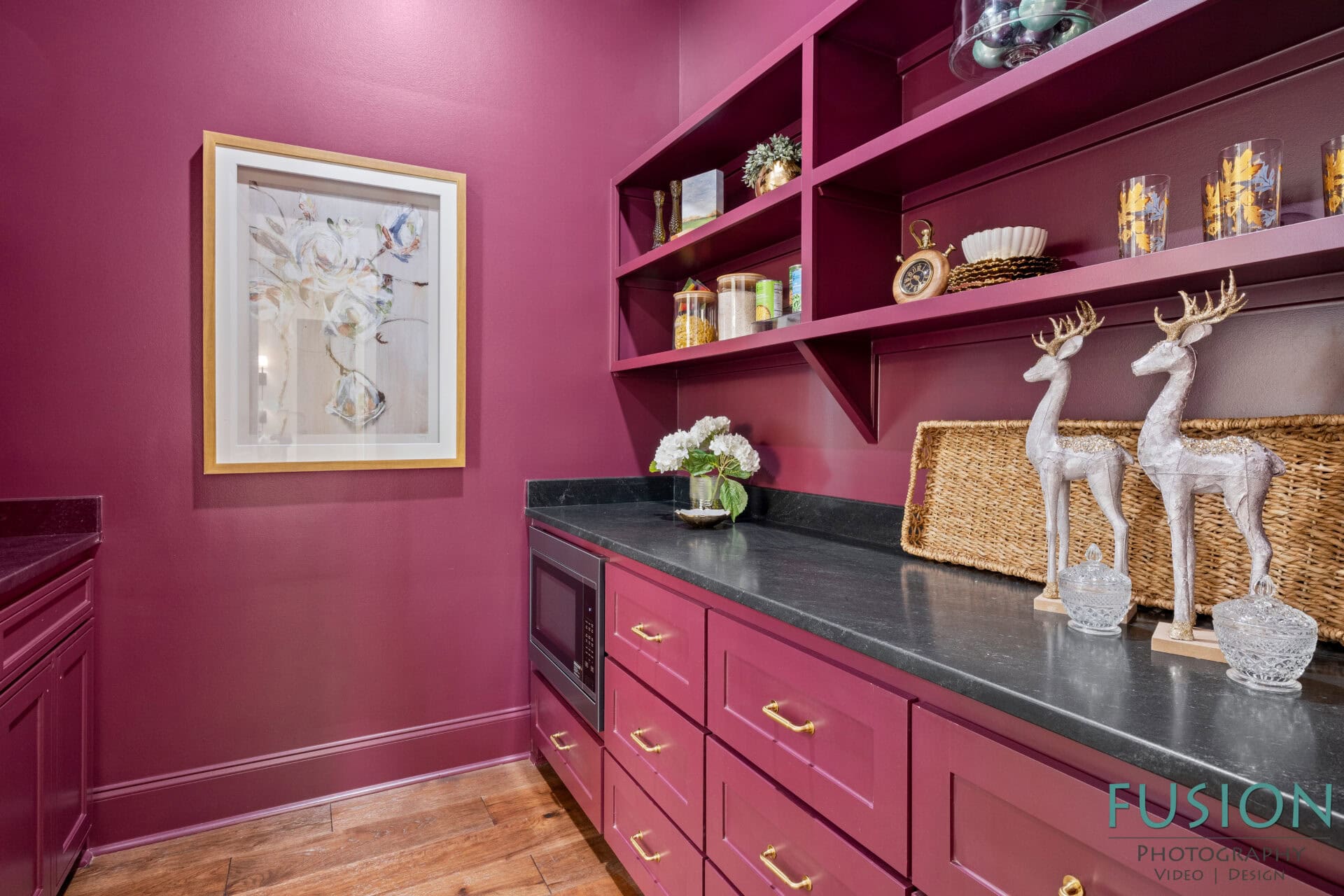 A pantry with deep magenta cabinets, black countertops, open shelves with decor and pantry items, a framed botanical artwork on the wall, and two decorative silver deer figurines on the counter.