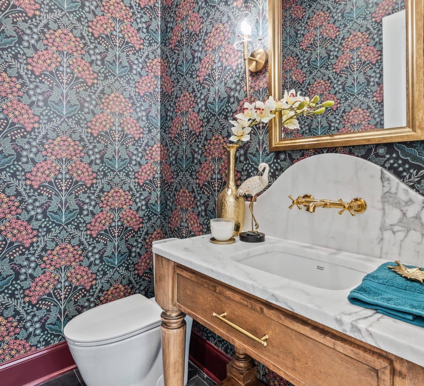 A small bathroom with floral wallpaper, a white toilet, a wooden vanity with a marble countertop, gold faucet, decorative crane figure, orchid, mirror, and a light fixture above the sink.