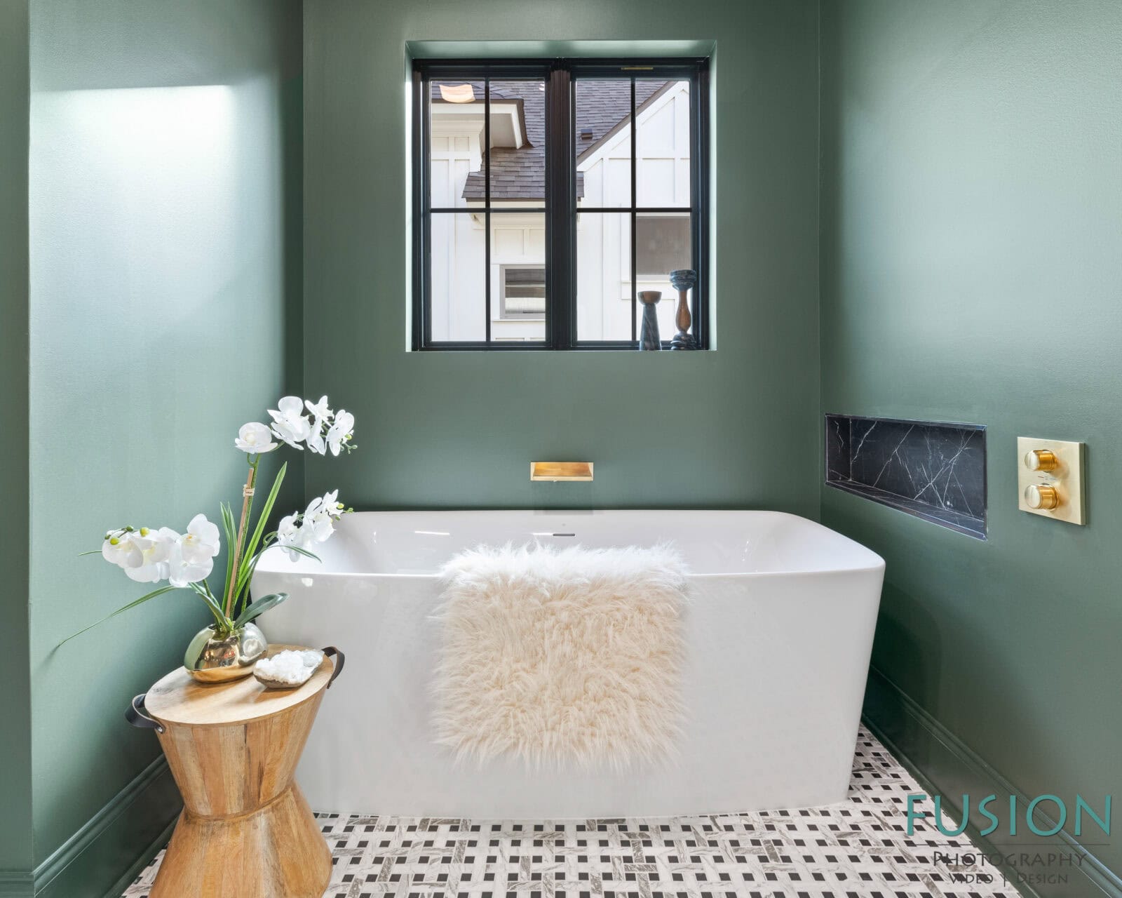A modern bathroom with green walls, a freestanding white bathtub with a fuzzy towel draped over the edge, a wooden stool with white flowers, patterned tile floor, and a window above the tub.