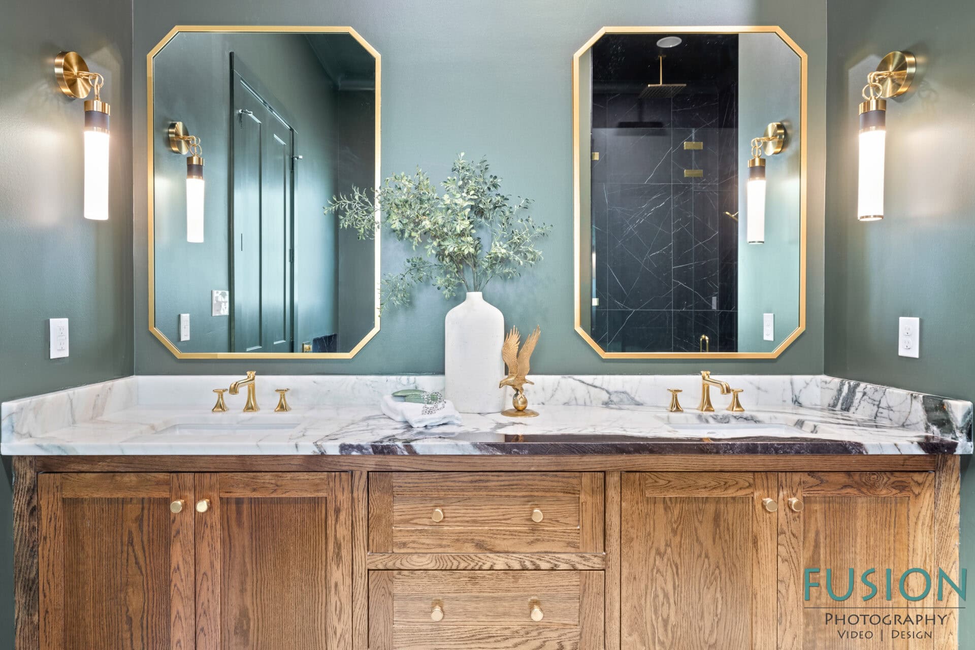 A bathroom features a double vanity with a marble countertop, wooden cabinets, gold faucets, two hexagonal mirrors, wall sconces, and a white vase with greenery. A shower with black tile is reflected in the mirrors.