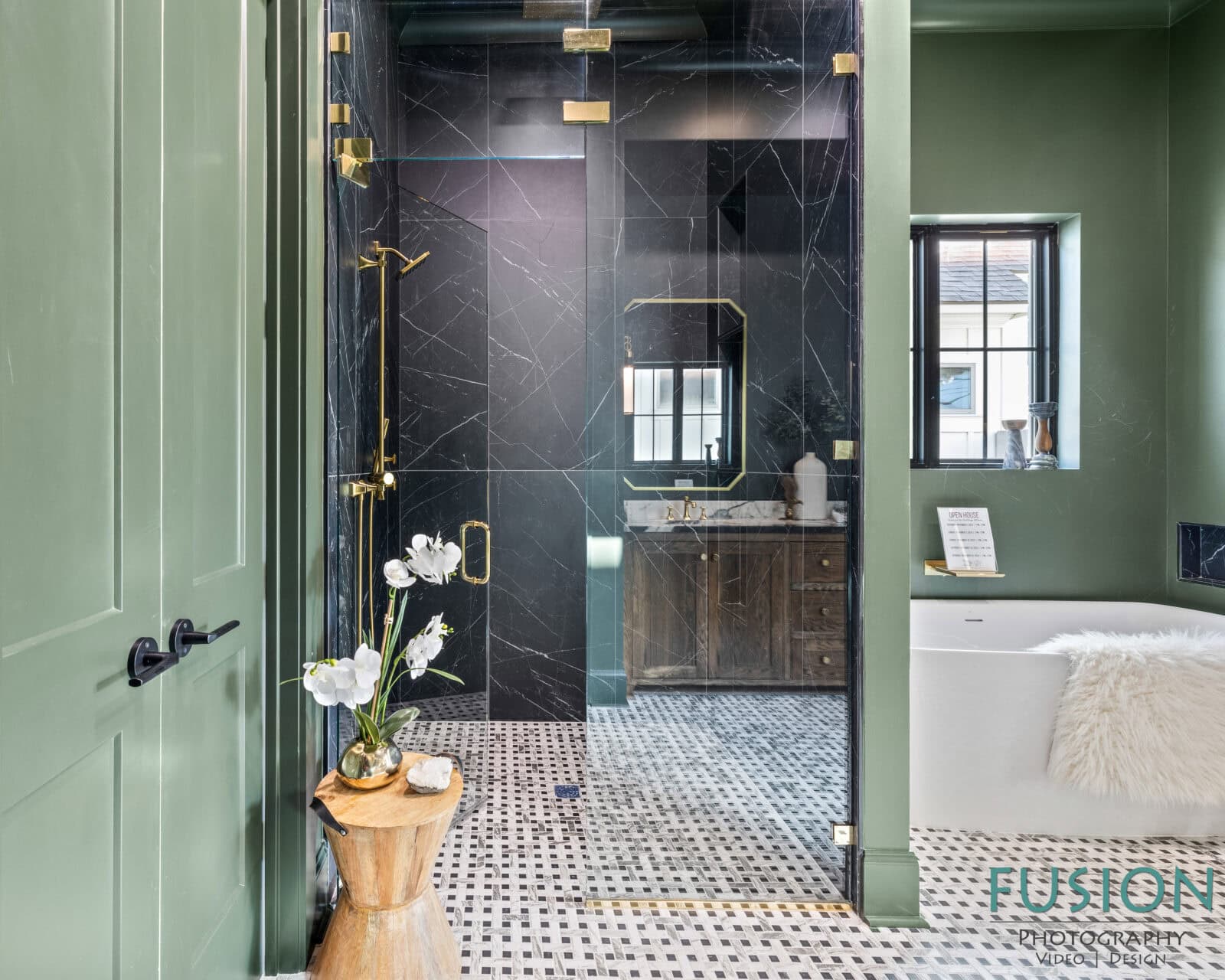 A modern bathroom features green walls, a black glass shower with gold hardware, a white bathtub, a wooden stool with a vase of white flowers, and a vanity with a large mirror. The floor has a black-and-white tile pattern.