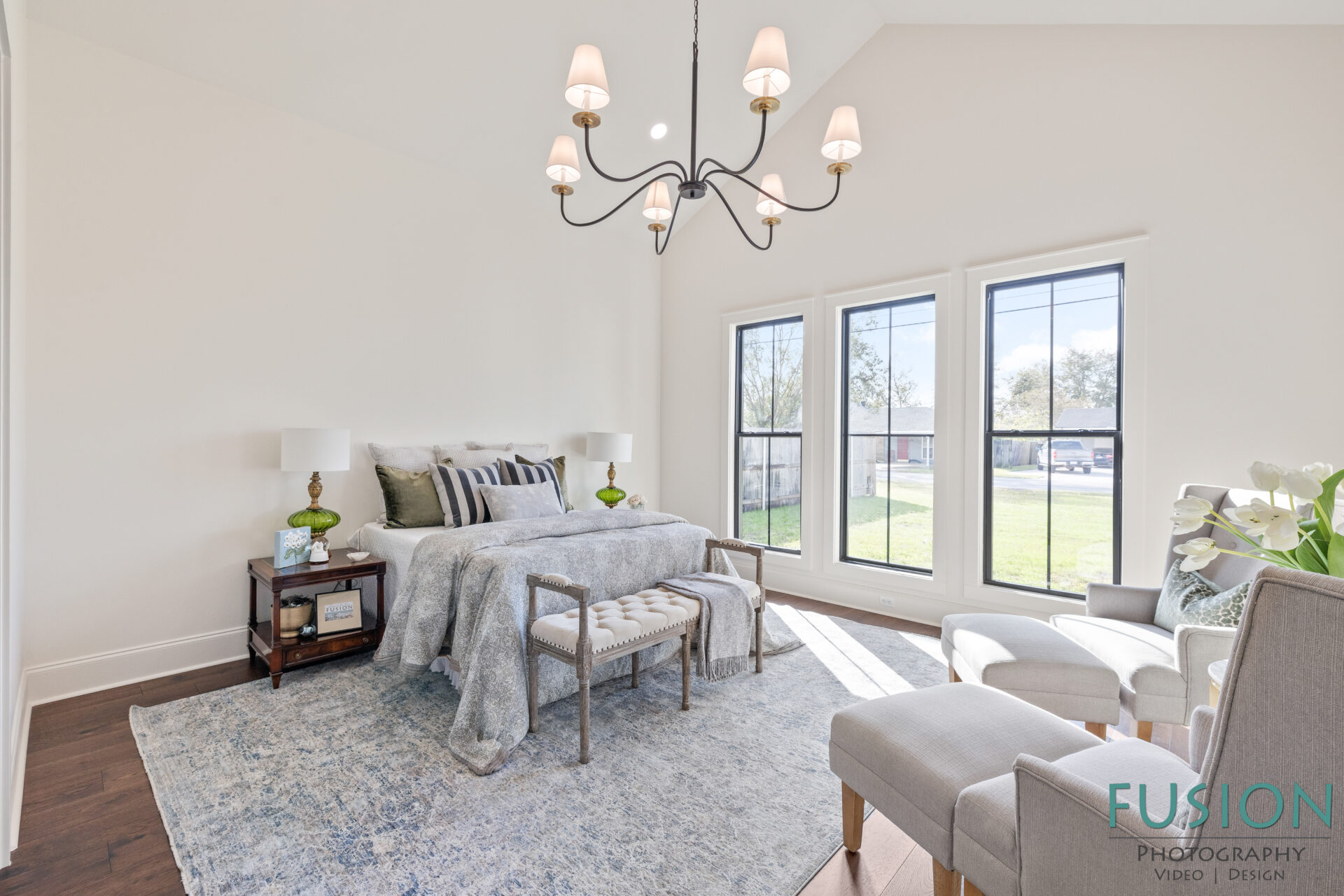 A bright bedroom with a large bed, two bedside tables with lamps, a cushioned bench at the foot of the bed, two armchairs, a blue area rug, three large windows, and a black chandelier.