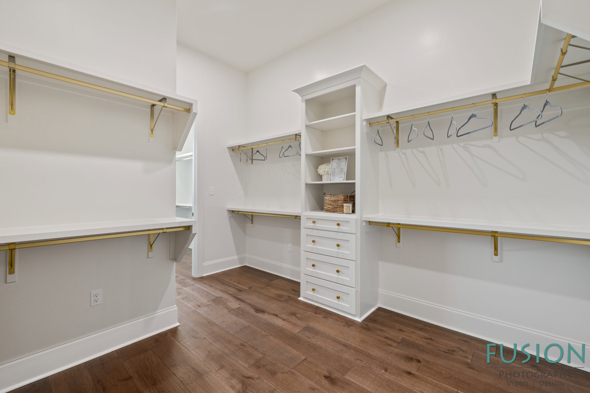 A spacious walk-in closet with white shelves, gold hanging rods, open cubbies, drawers with silver knobs, and several empty hangers. The floor is dark wood and the walls are white.