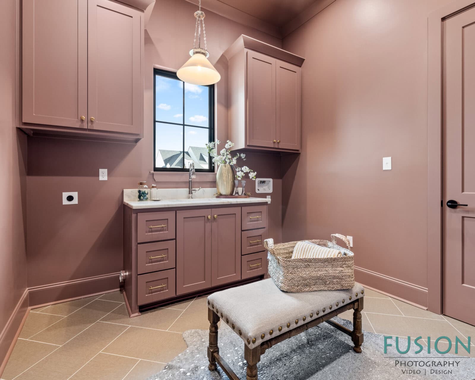 A mauve-colored laundry room features a sink with cabinets above and below, a window, a pendant light, a bench with a basket on top, and beige tile flooring. The Fusion Photography logo appears in the bottom right corner.