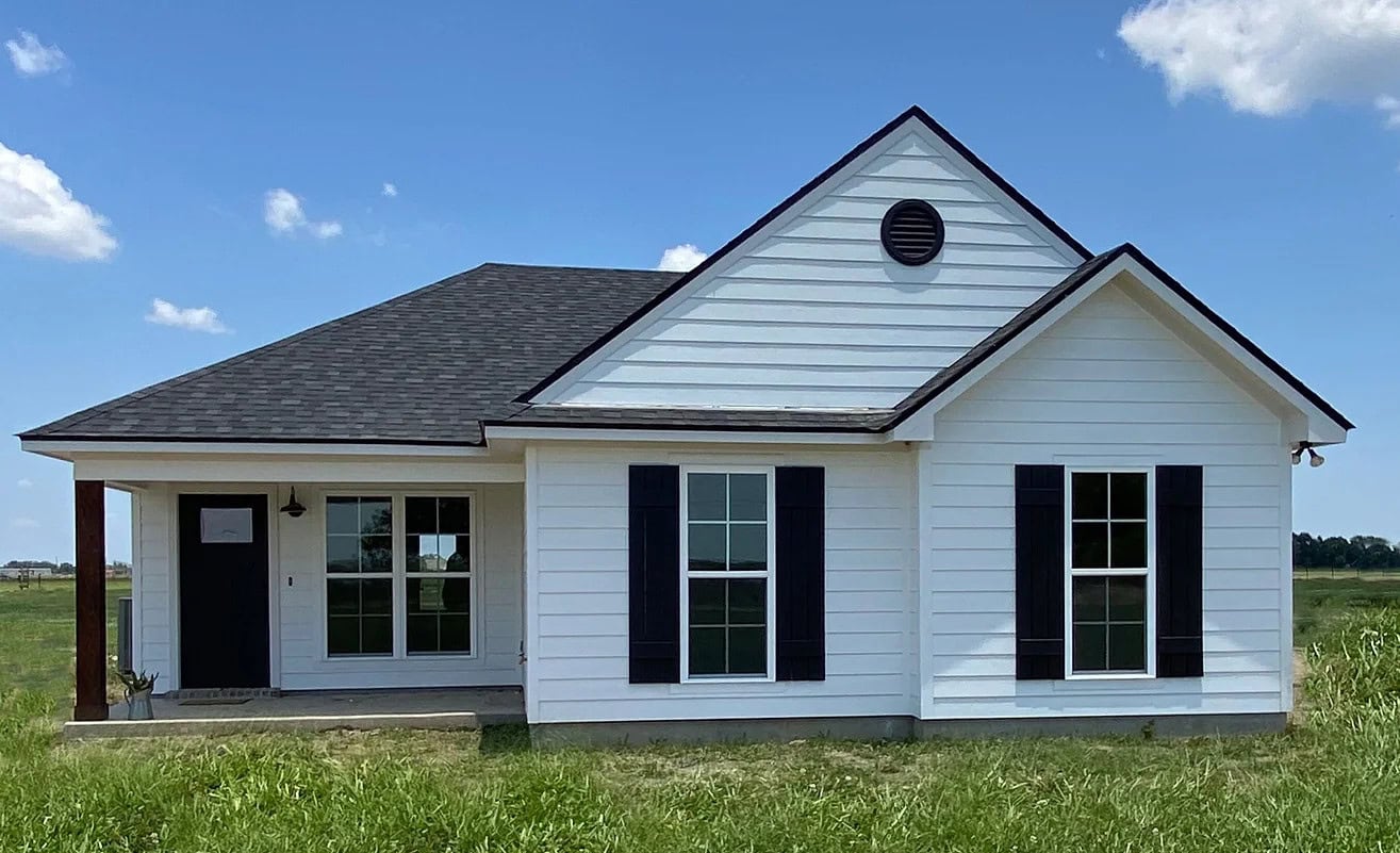 A small, single-story white house with black shutters and a dark roof sits in a grassy field under a blue sky with a few clouds. The house has two sets of windows and a covered entryway.