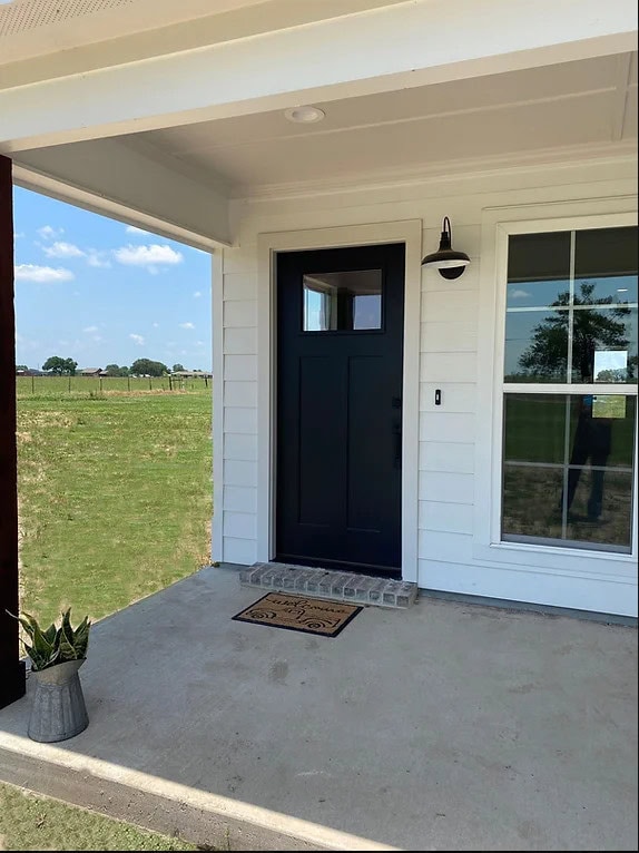 A black front door with a window sits on a white house with siding. A welcome mat and potted plant are on the concrete porch. A single outdoor light is mounted beside the door. A field and blue sky are in the background.
