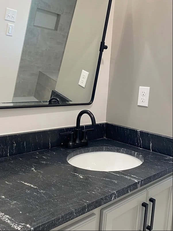 A bathroom countertop with a dark stone surface, an oval white sink, a black faucet, and a large rectangular mirror. There are electrical outlets and light gray walls in the background.