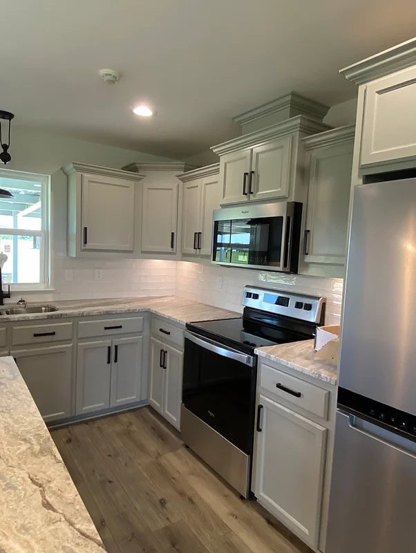 Modern kitchen with light gray cabinets, stainless steel appliances, electric stove, built-in microwave, marble-look countertops, white tile backsplash, and wood-look flooring. Window above the sink provides natural light.