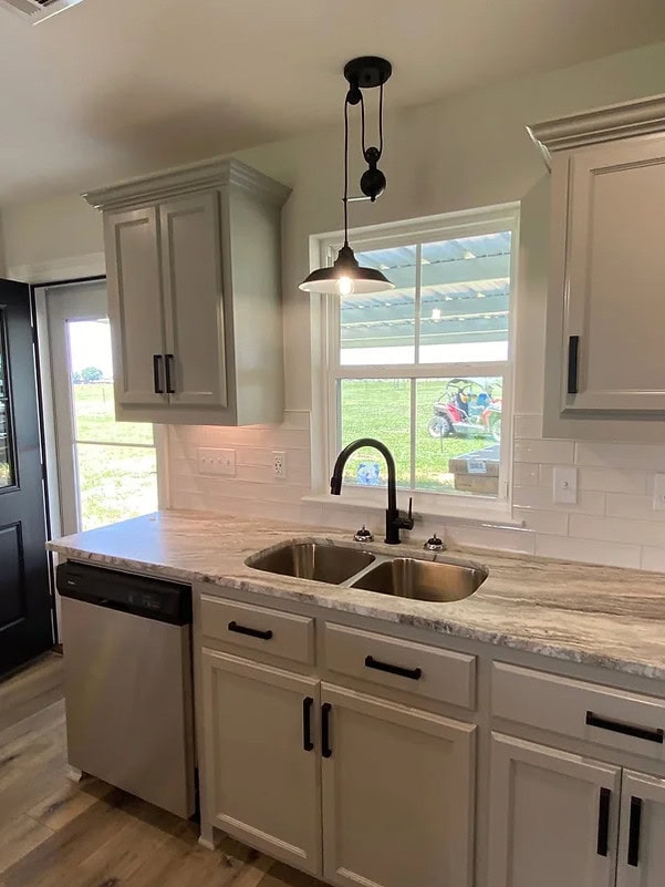 A kitchen countertop with a double sink, stainless steel dishwasher, light gray cabinets, black handles, a pendant light above the sink, and a window showing a green lawn with a toy car outside.