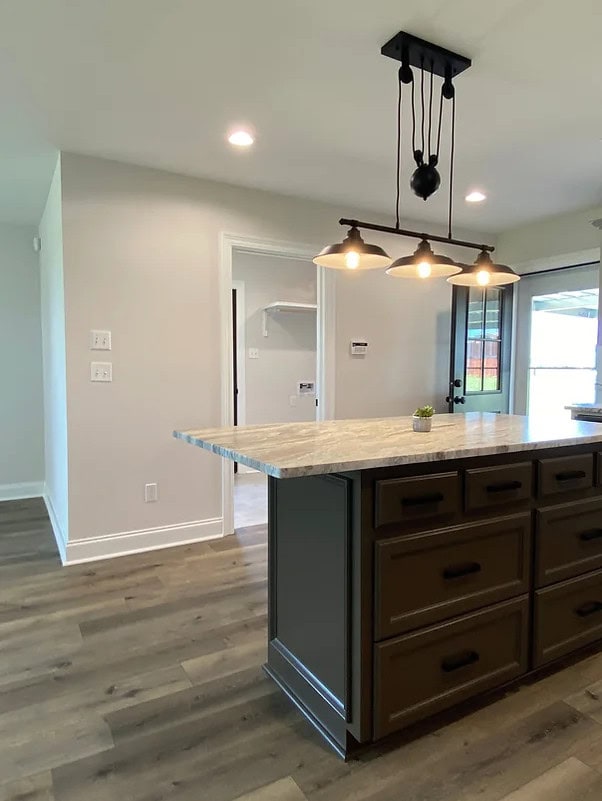 A modern kitchen with a large island featuring a marble countertop, dark brown drawers, a small plant, and an overhead black light fixture with three bulbs. The floor is wood, and walls are light-colored.