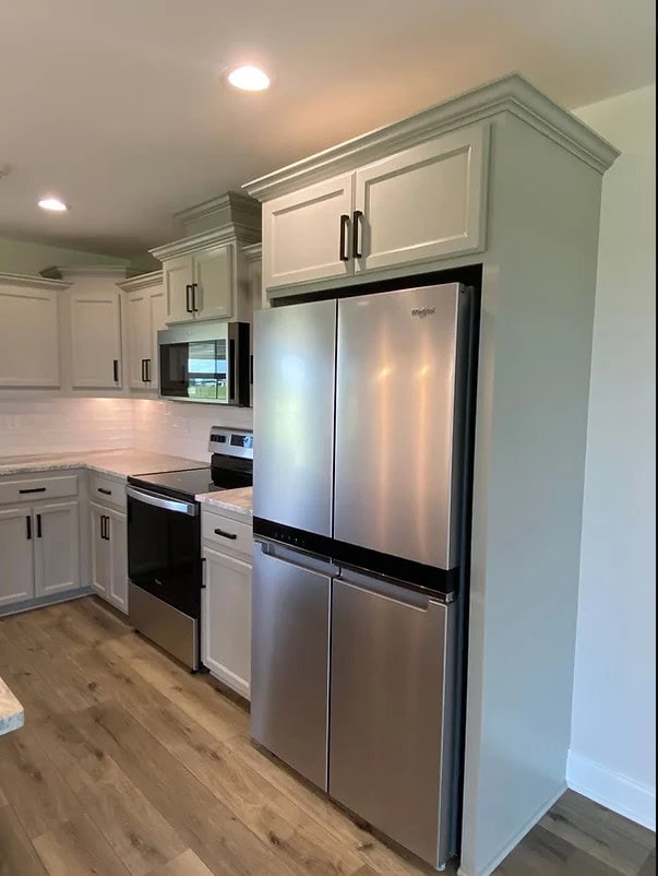 A modern kitchen with light gray cabinets, stainless steel appliances, a French-door refrigerator, an electric stove, a microwave, and light wood flooring. Recessed lighting is installed in the ceiling.