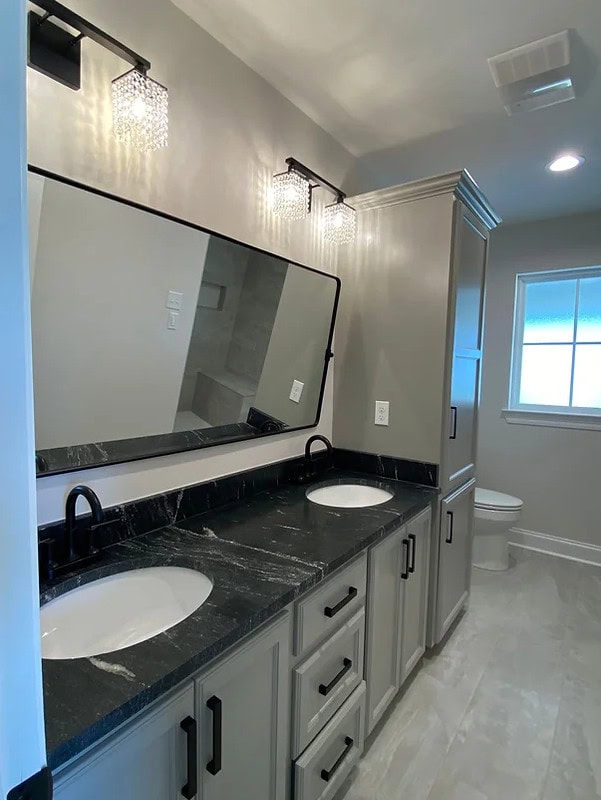 A modern bathroom with a double sink vanity featuring black countertops, gray cabinets, a large rectangular mirror, two wall-mounted light fixtures, and a toilet near a frosted window.