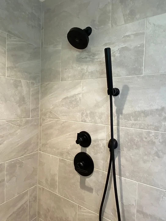 A tiled shower wall with a black showerhead, a handheld shower wand on a vertical bar, and two black control knobs mounted below the showerhead.