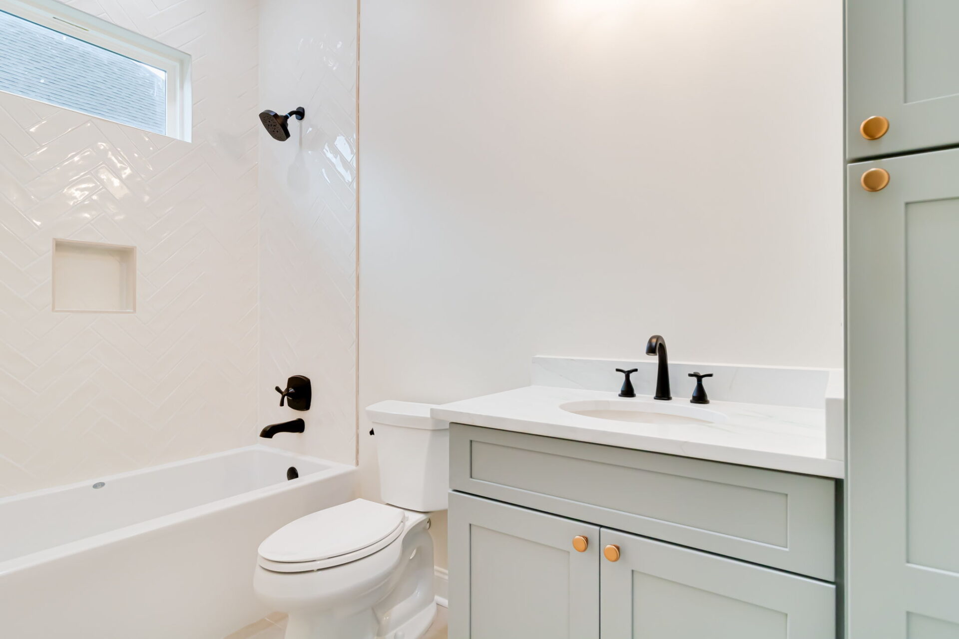 A modern bathroom with a white bathtub and shower, a white toilet, and a light gray vanity with a white countertop and black fixtures. The shower wall has a herringbone tile pattern and a built-in niche.