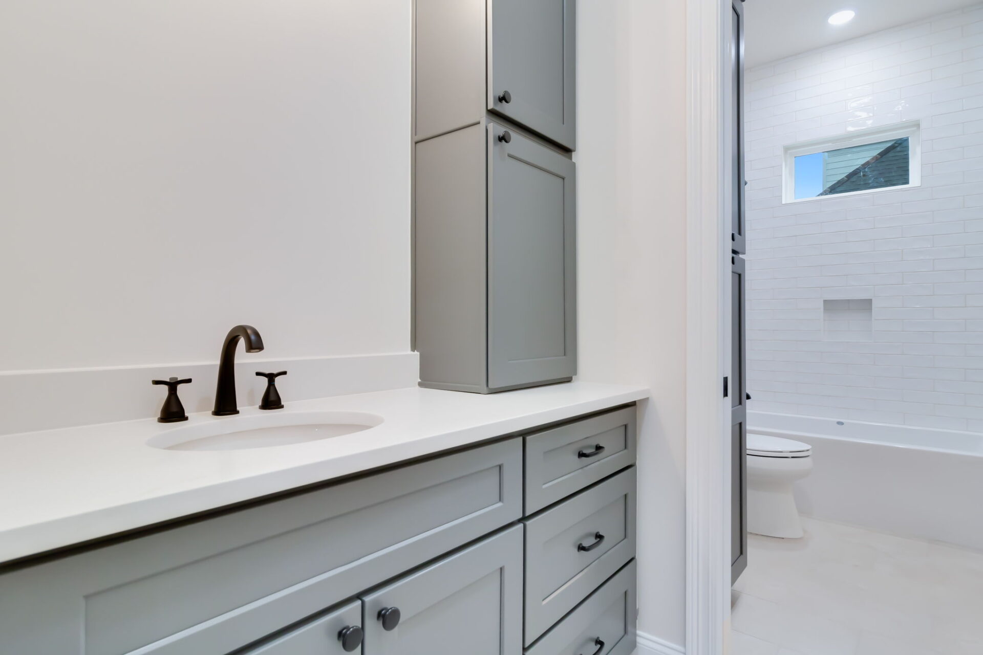 A modern bathroom with light gray cabinets, a white countertop, black faucet, and a built-in sink. In the background, there is a white bathtub, toilet, and a window above tiled walls.