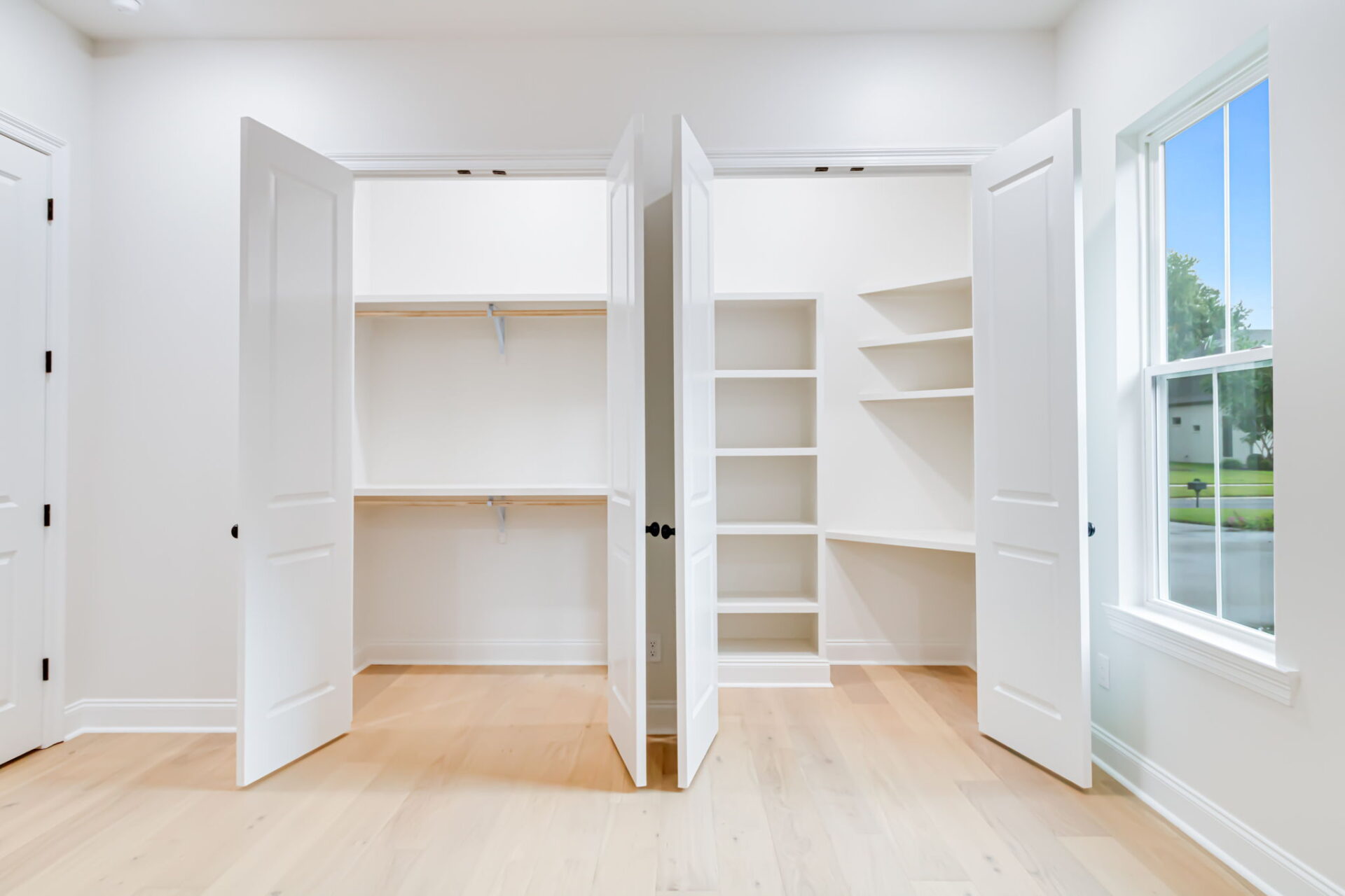 A bright, empty closet with double doors, open to reveal shelves, angled corner shelves, and hanging rods. The closet has light wood floors and white walls. A window to the right shows a green lawn outside.