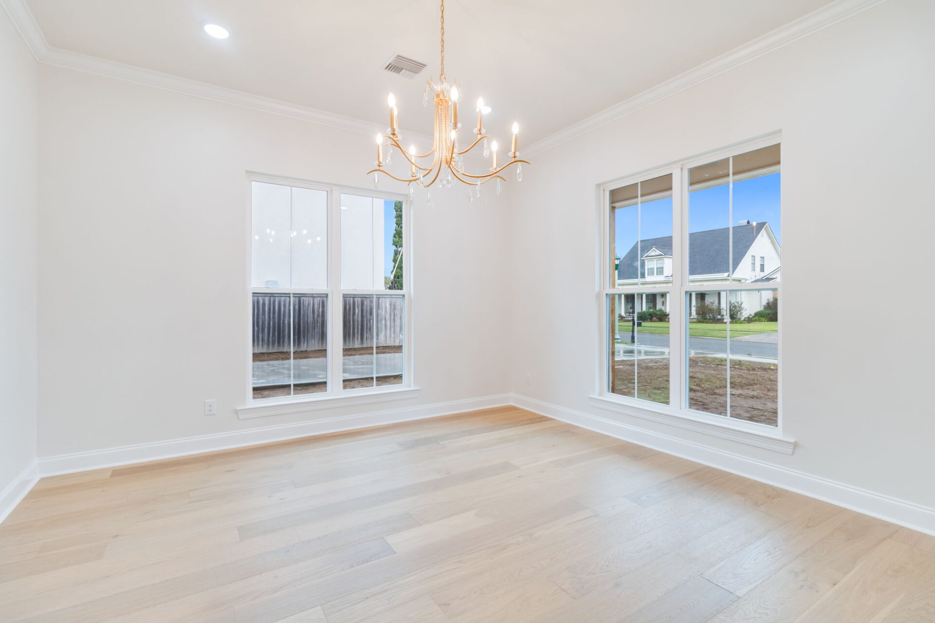 A bright, empty room with light wood flooring, white walls, two large windows, and a gold chandelier hanging from the ceiling. The windows show a view of a fenced yard and a house across the street.