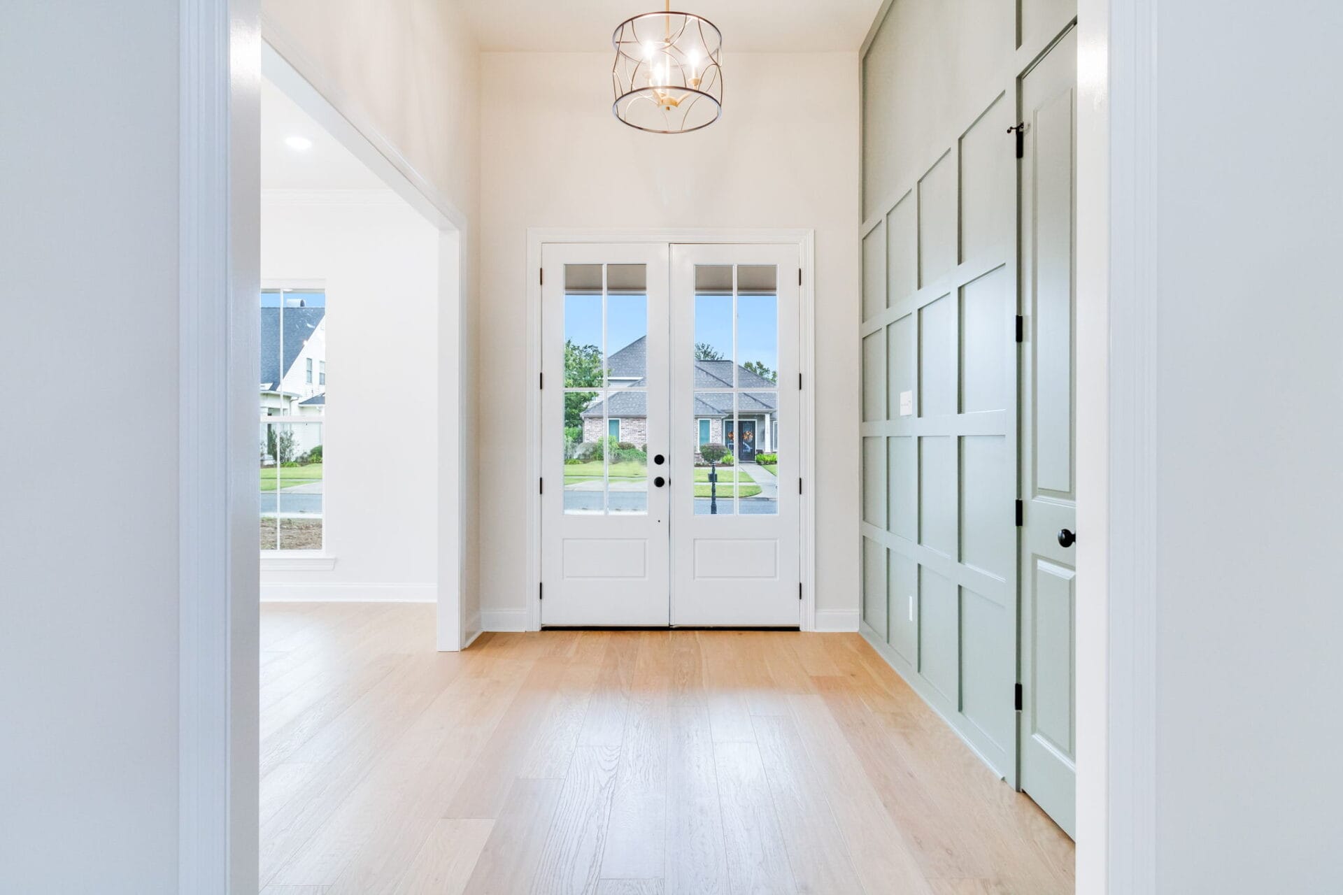 Bright entryway with light wood flooring, white walls, a green accent wall with paneling on the right, double glass doors, and a modern ceiling light fixture. Outdoor suburban homes are visible through the doors.
