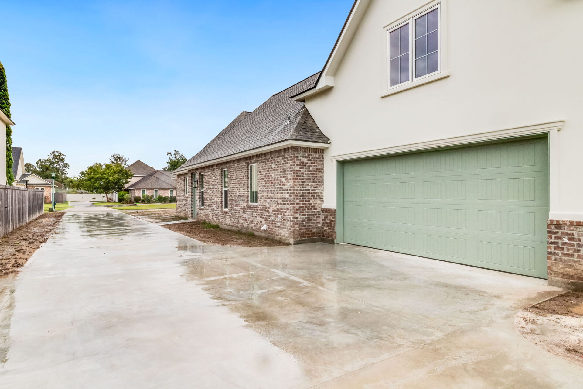 A concrete driveway leads to a two-car garage with light green doors on a brick and stucco house. The sky is clear and neighboring houses are visible in the background.