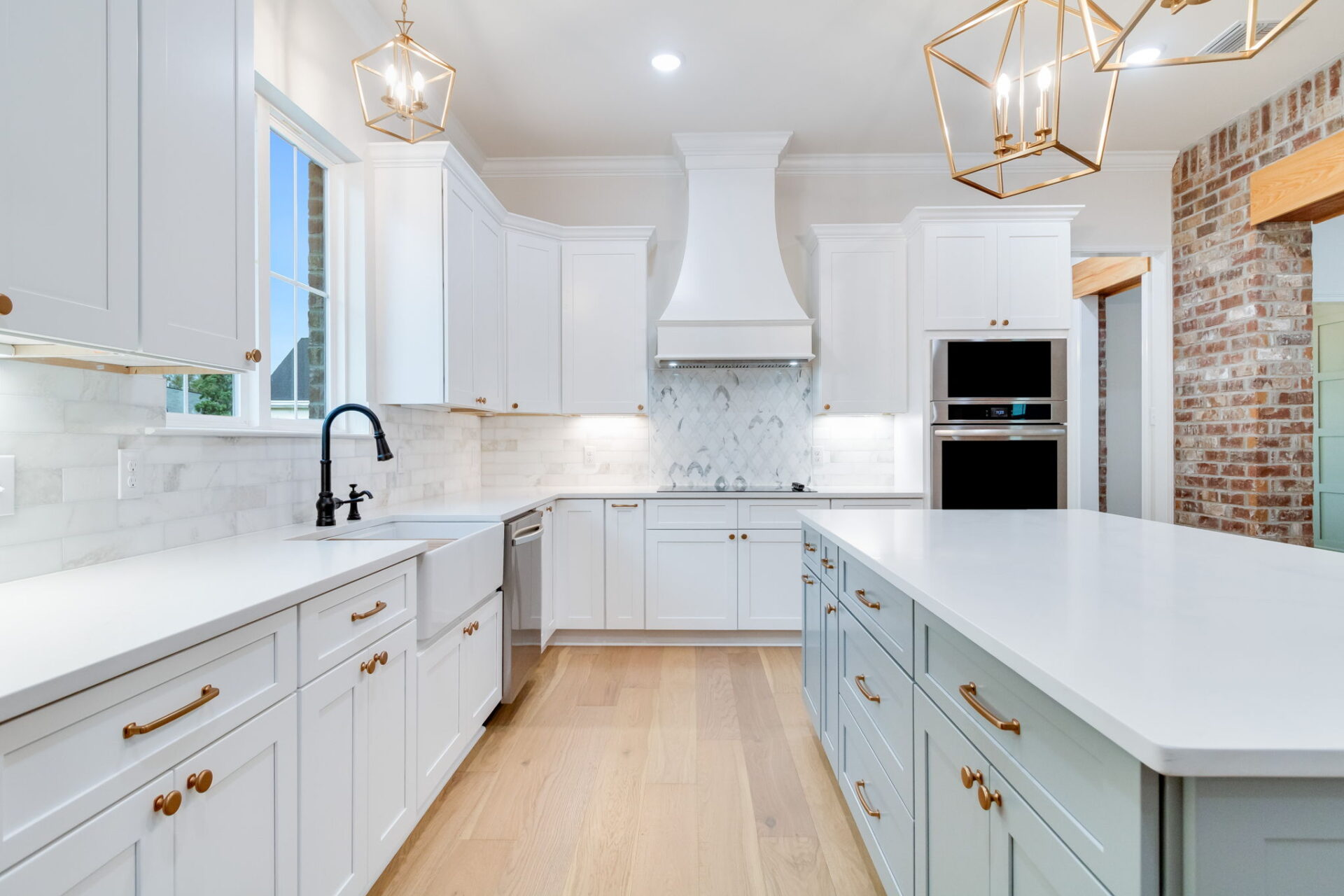 Spacious modern kitchen with white cabinets, gold hardware, marble backsplash, large island, light wood flooring, stainless steel appliances, farmhouse sink, and geometric pendant lights. Brick wall is visible on the right.