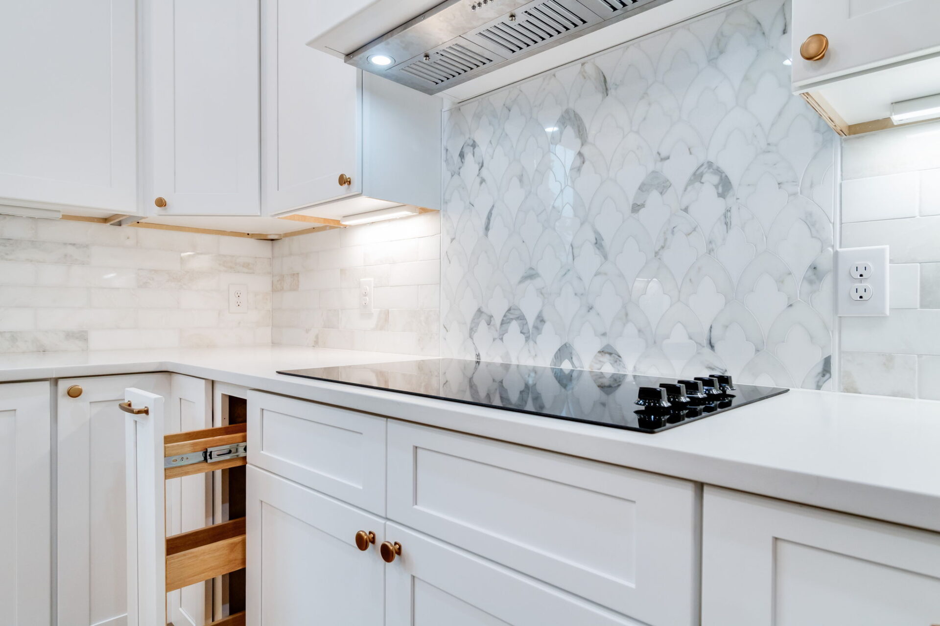 A modern kitchen with white cabinets, marble tile backsplash, a black glass electric cooktop, pull-out storage drawer, and under-cabinet lighting. The counters are clean and the wall outlets are visible.