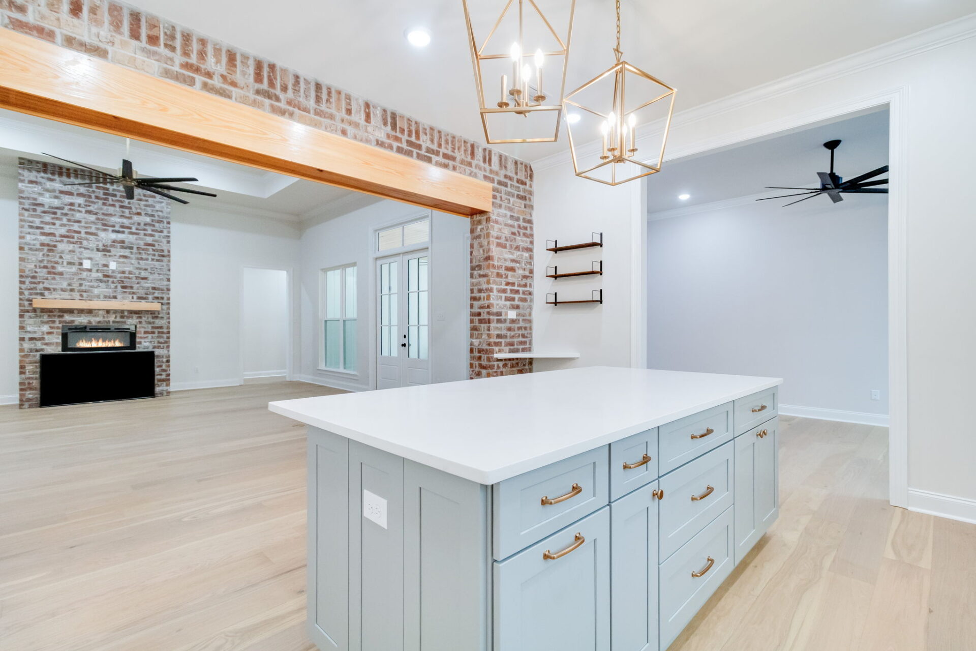 A modern open-concept kitchen and living area with light wood floors, a large island with blue cabinets and gold hardware, exposed brick wall, fireplace, and two ceiling fans.