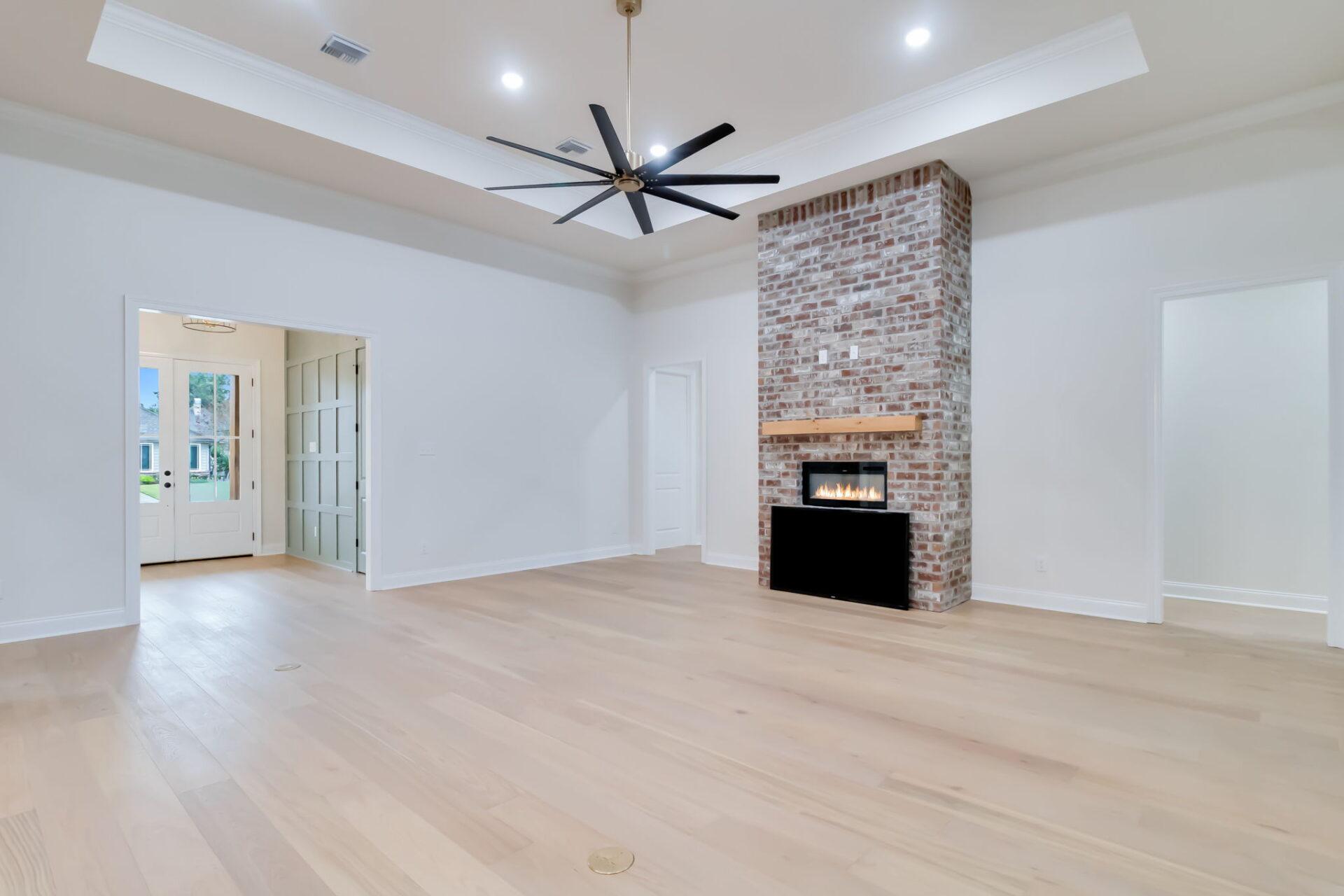 A spacious, empty living room with light wood floors, white walls, a brick fireplace with a wooden mantel, recessed ceiling lights, and a large black ceiling fan. Two doors and a doorway lead to other rooms.