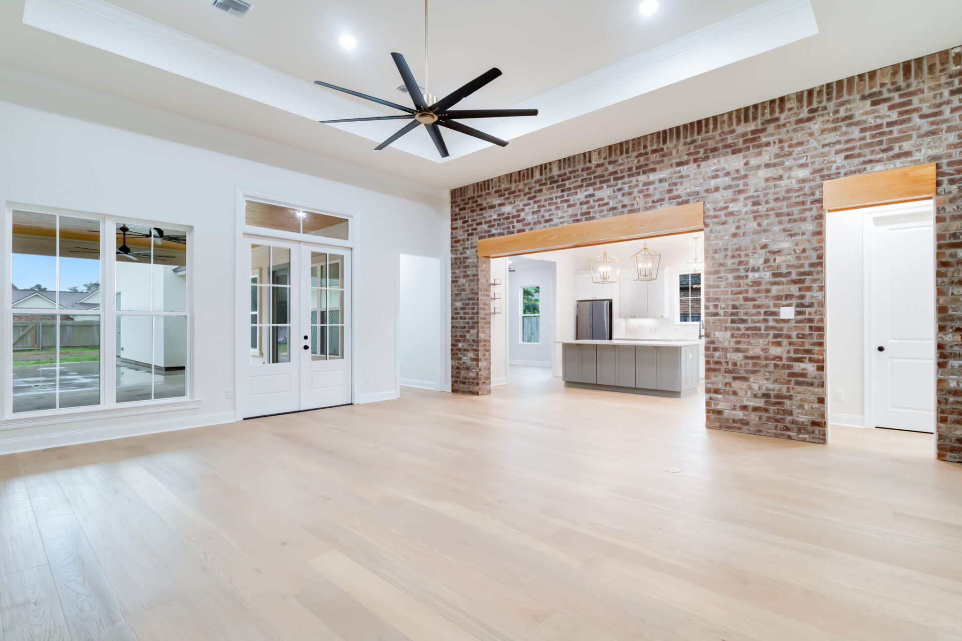 A spacious, empty living area with light wood floors, brick accent wall, large ceiling fan, and open view into a modern kitchen with pendant lights. French doors and windows open to an outdoor space.