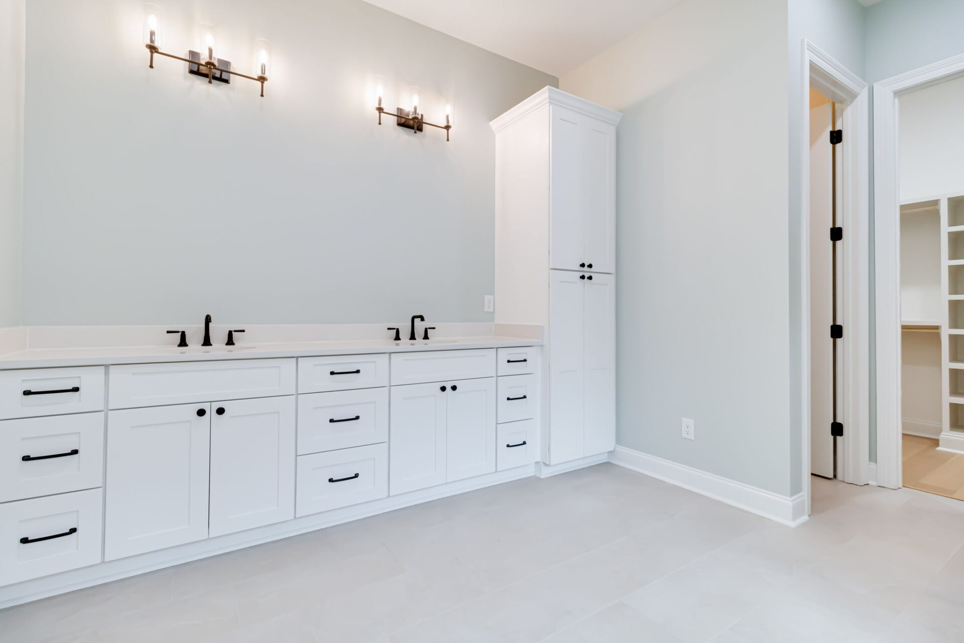 A modern bathroom with white double vanity cabinets, two sinks with black fixtures, wall-mounted light fixtures above, and a tall storage cabinet. The room has light-colored walls and floor tiles.
