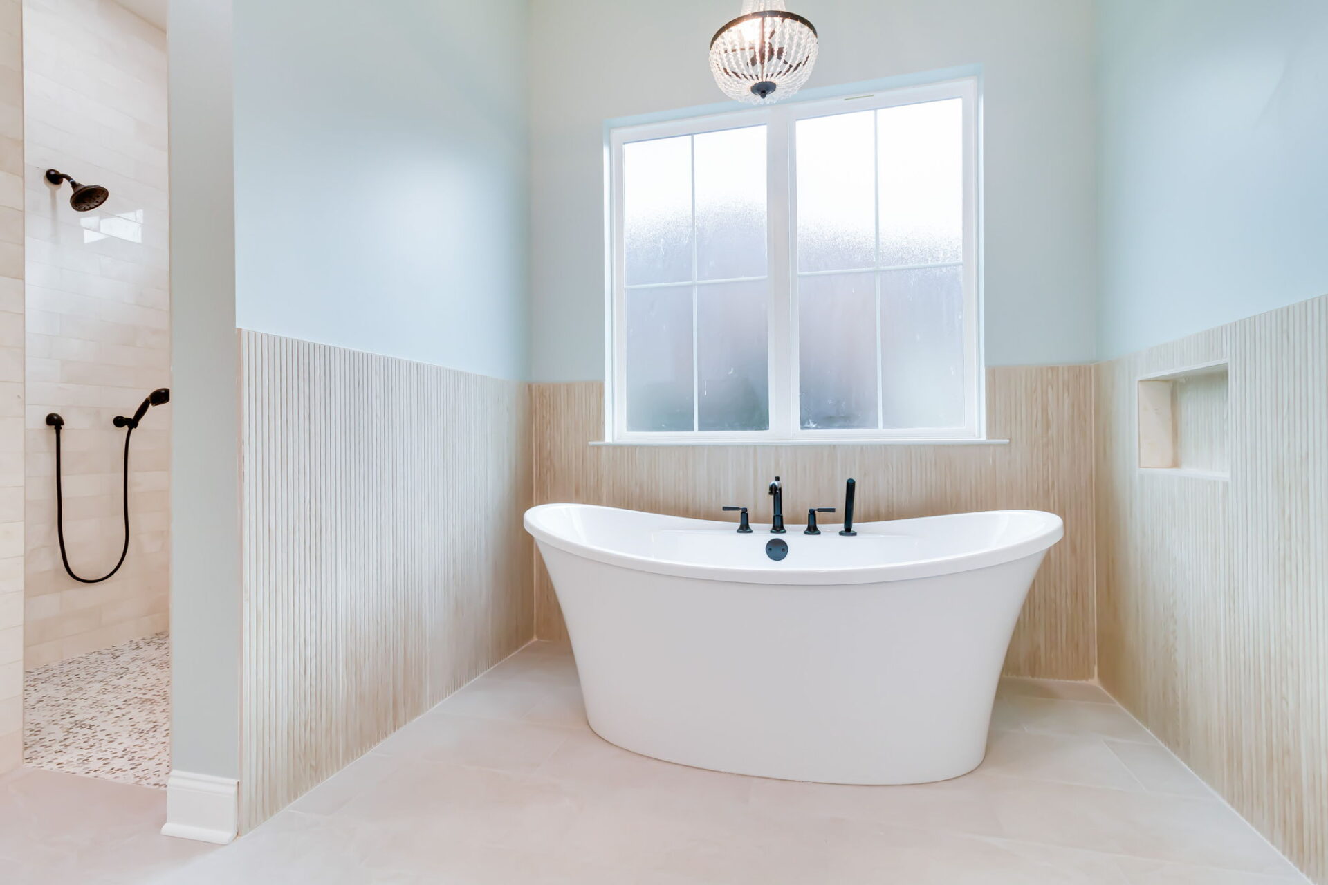 A modern bathroom with a freestanding white bathtub, matte black fixtures, a large frosted window, light-colored walls and tiles, and a walk-in shower with a glass door and black showerhead in the background.