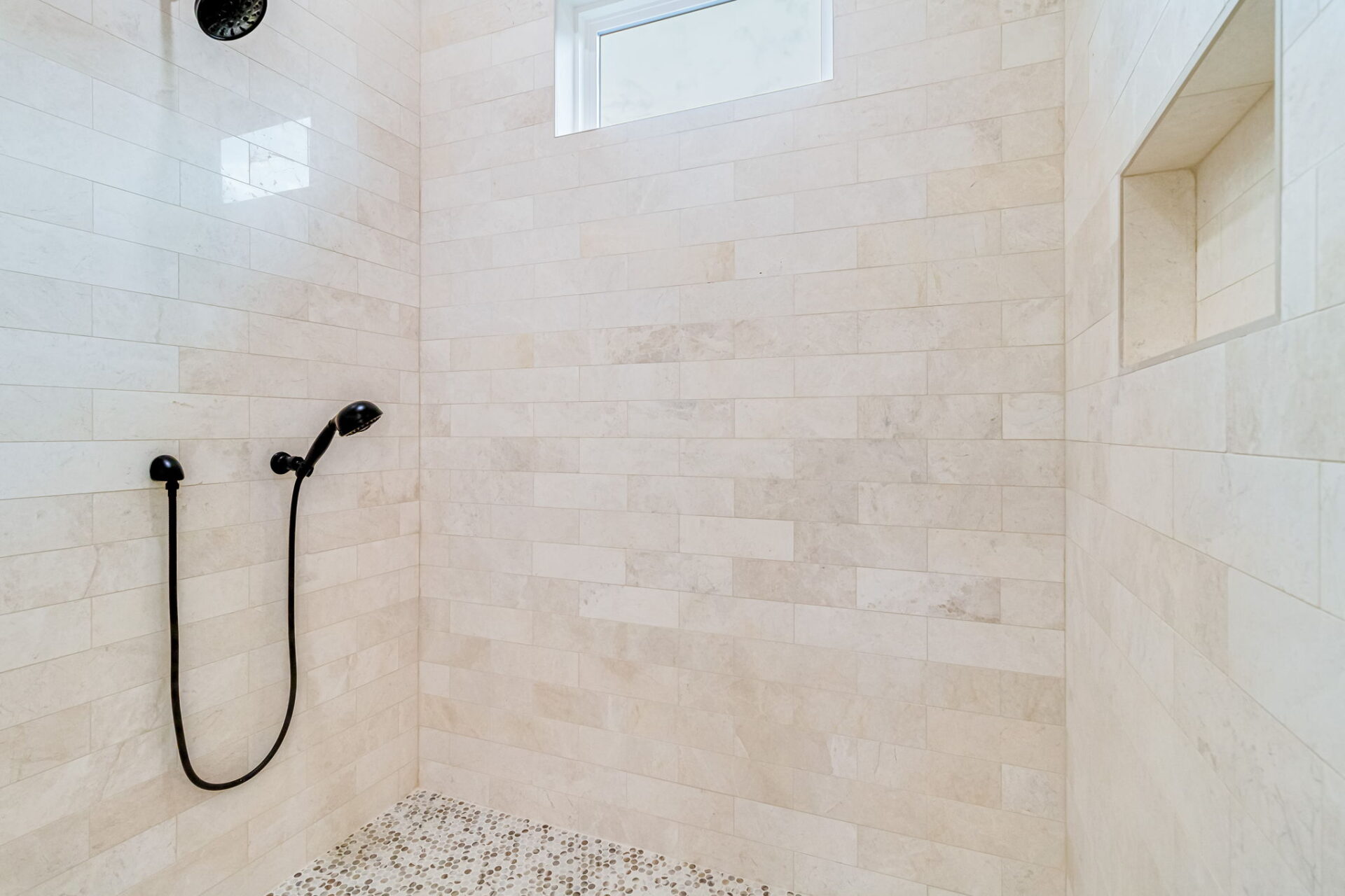 Walk-in shower with beige rectangular tiles, a built-in wall niche, a small high window, a black handheld showerhead, and a mosaic tile floor.