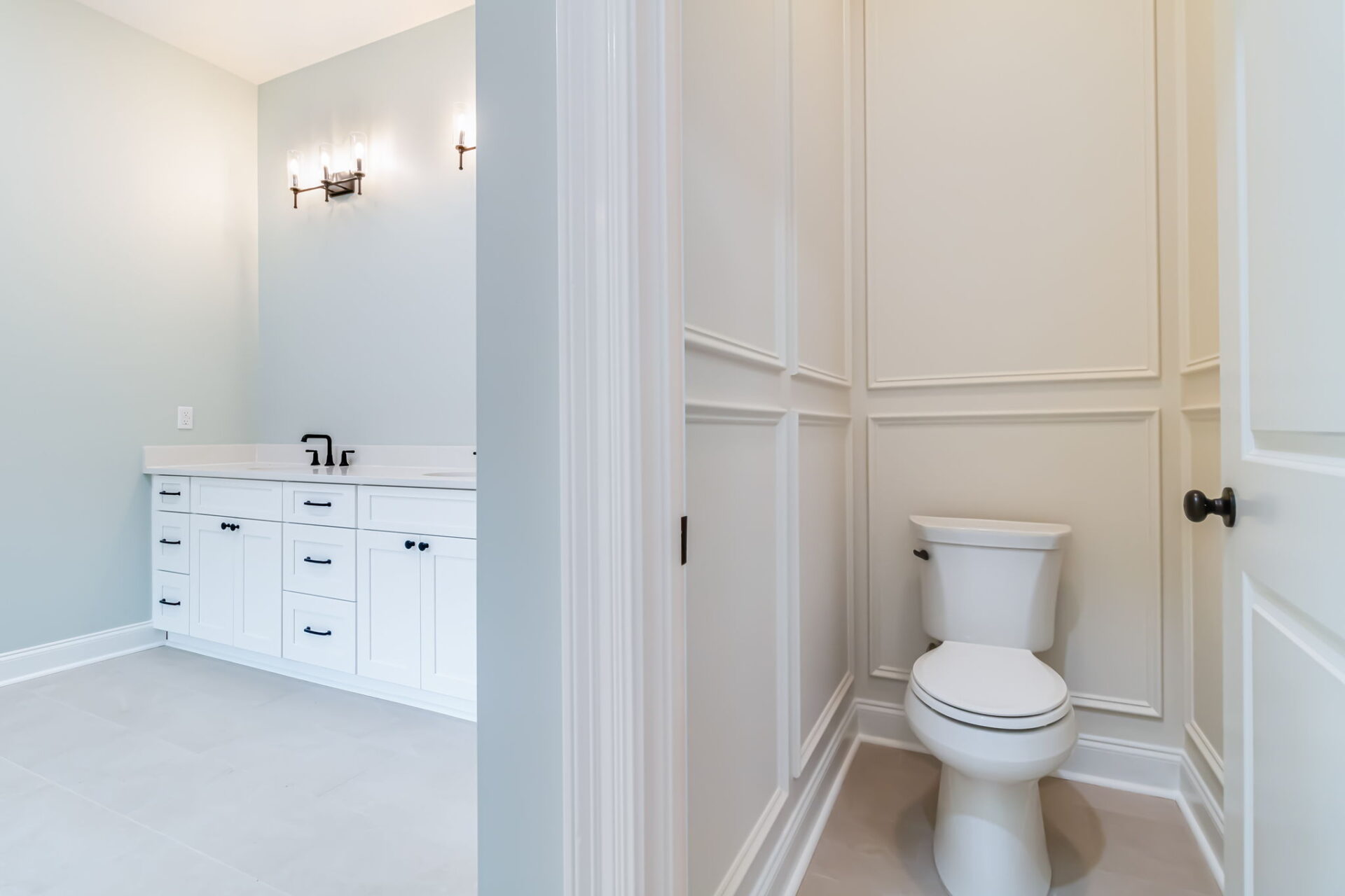 A modern bathroom with white cabinets, double sinks, and wall-mounted light fixtures is shown next to a separate enclosed area containing a white toilet. The room features light-colored walls and flooring.