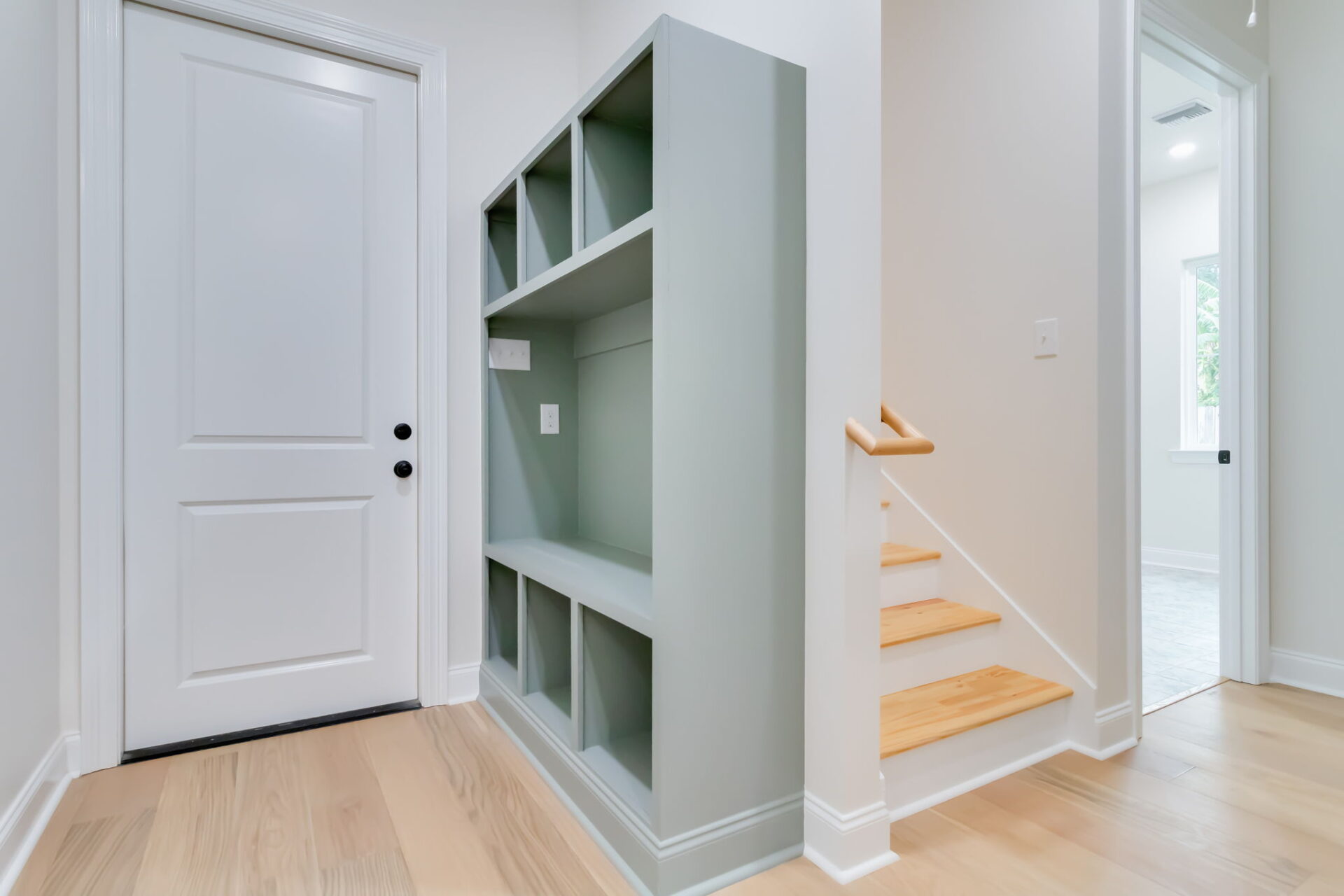 A modern hallway with light wood floors, a white door, a built-in light green storage shelf, and a small wooden staircase with a simple handrail leading up to another room.