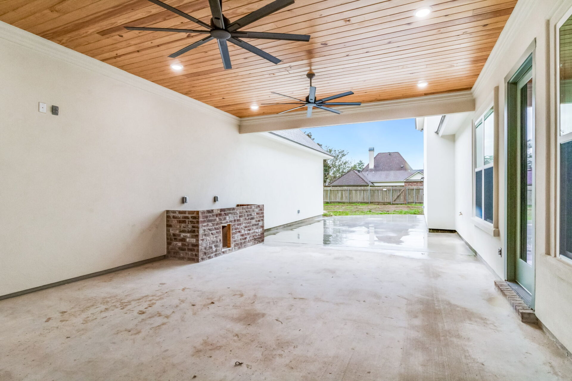 Covered outdoor patio with a wood-paneled ceiling, recessed lighting, two ceiling fans, a small brick fireplace on the left wall, and large windows on the right overlooking a fenced backyard.