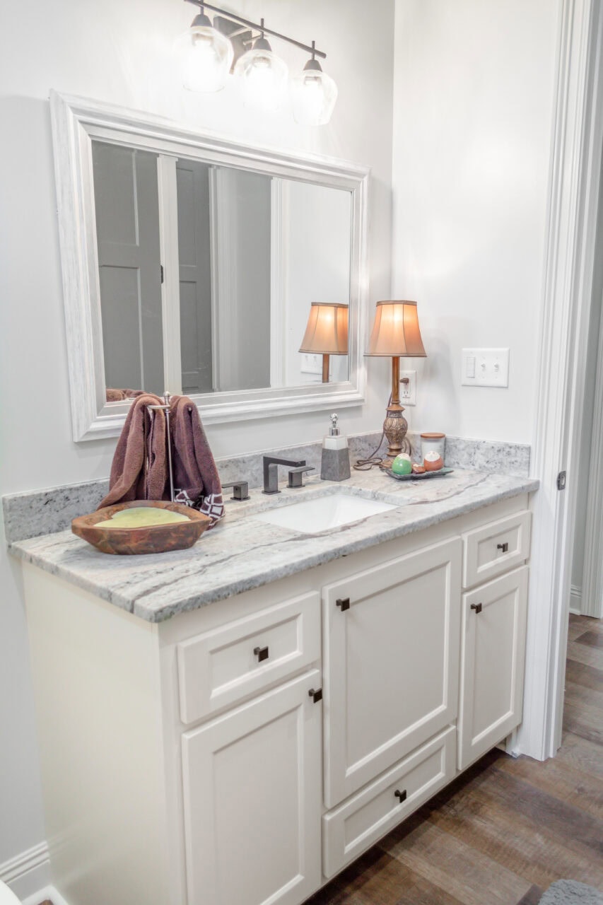 A bathroom vanity with a white countertop, sink, faucet, and cabinets. A mirror hangs above, with three vanity lights. A brown lamp, a brown towel, a bowl, and toiletries sit on the counter. The floor is wooden.