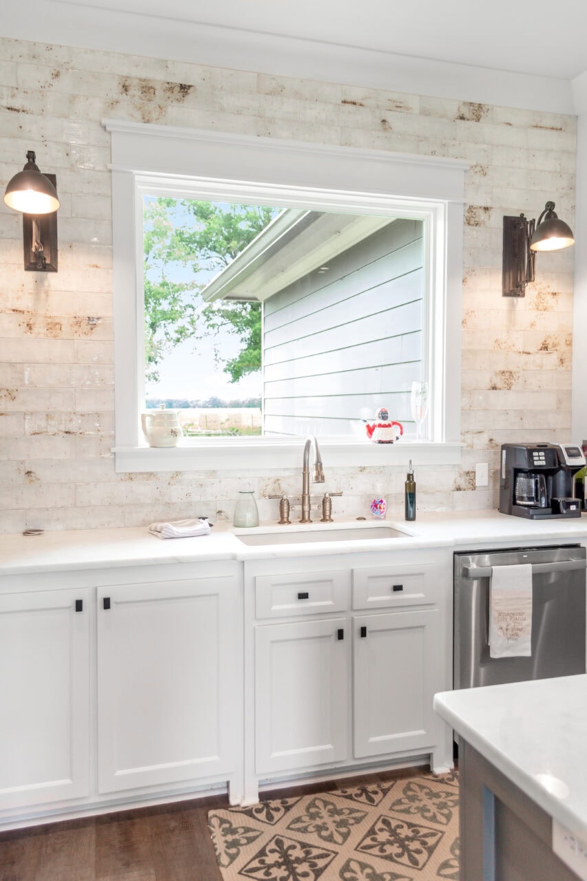 A bright kitchen features white cabinets, a marble countertop, a large window with a view outside, stainless steel sink, coffee maker, patterned floor mat, and rustic wall sconces.