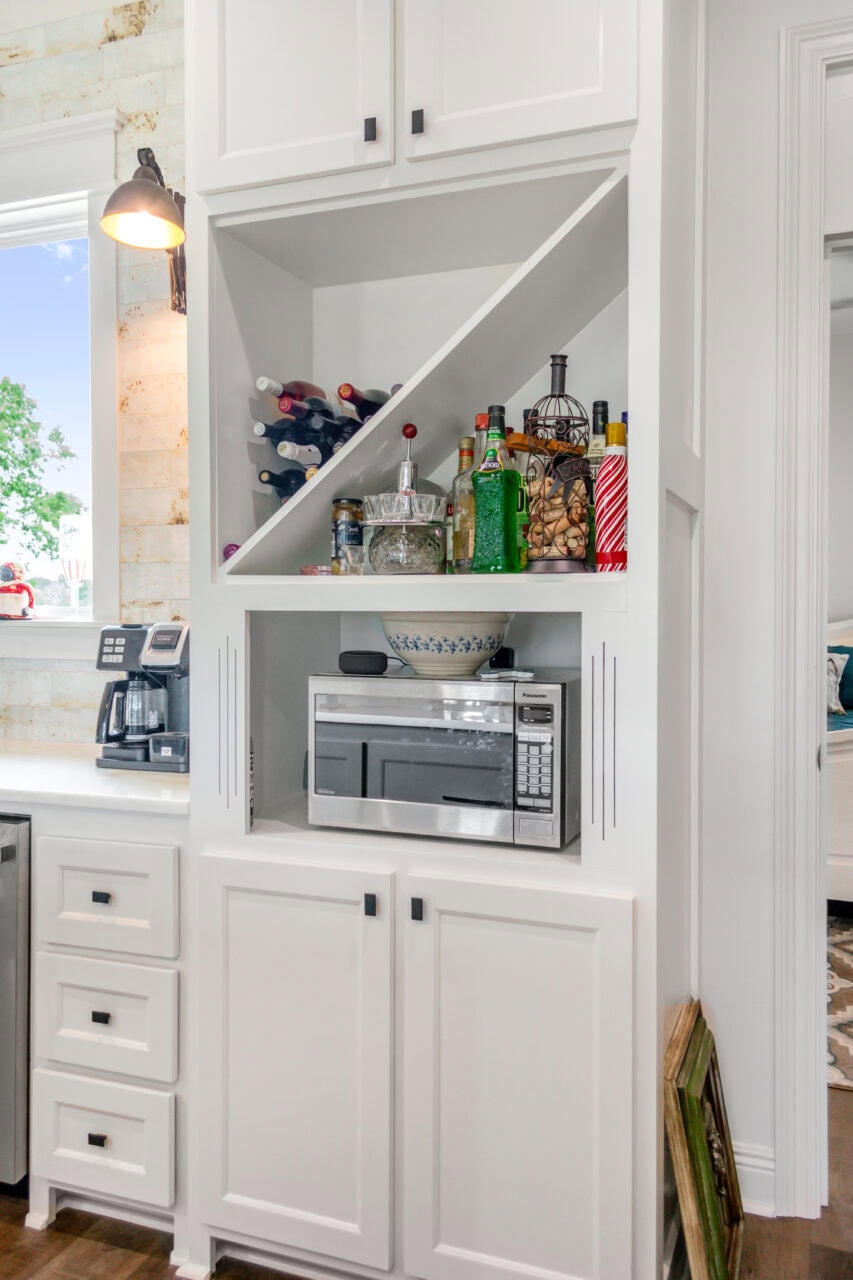A white built-in kitchen cabinet with diagonal shelves holding wine bottles and liquor, a middle shelf with a bowl, snacks, and drinks, and a lower shelf with a microwave. The counter holds kitchen appliances.