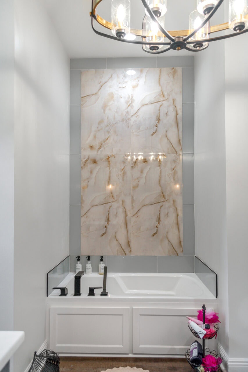 A white bathroom features a rectangular bathtub with black fixtures, toiletries on the ledge, a large marble-patterned tile accent on the wall, and a modern chandelier reflected above the tub.
