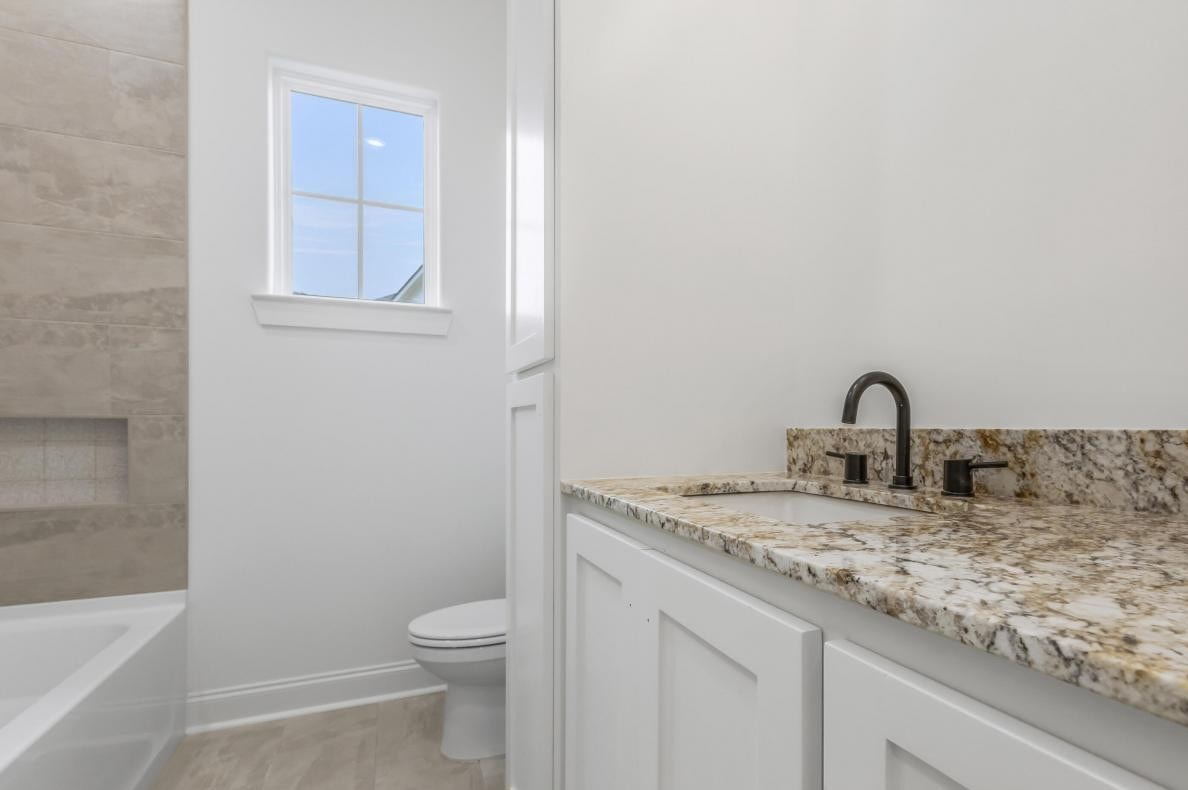 A bathroom with a granite countertop, white cabinets, an undermount sink with a black faucet, a toilet, a window, and a bathtub with beige tile walls.