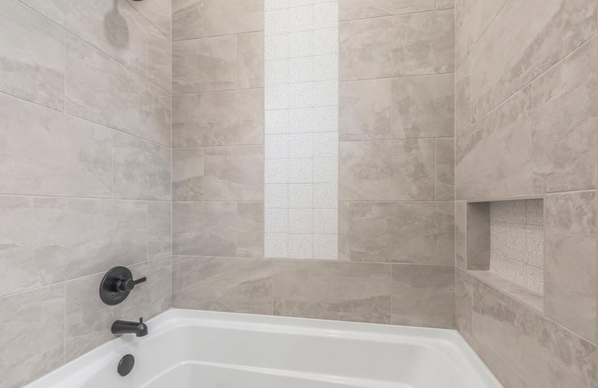 A white bathtub with light beige rectangular tiles on the walls, featuring a vertical strip of smaller white tiles and a built-in recessed shelf. Black fixtures for the faucet, handle, and showerhead are visible.