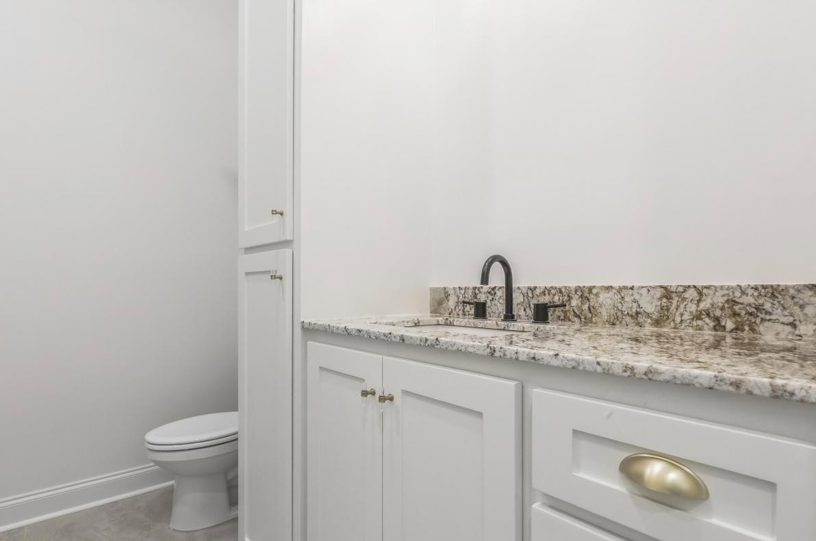 A bathroom with white cabinets, a granite countertop, a black faucet, and a toilet in the background against a white wall.