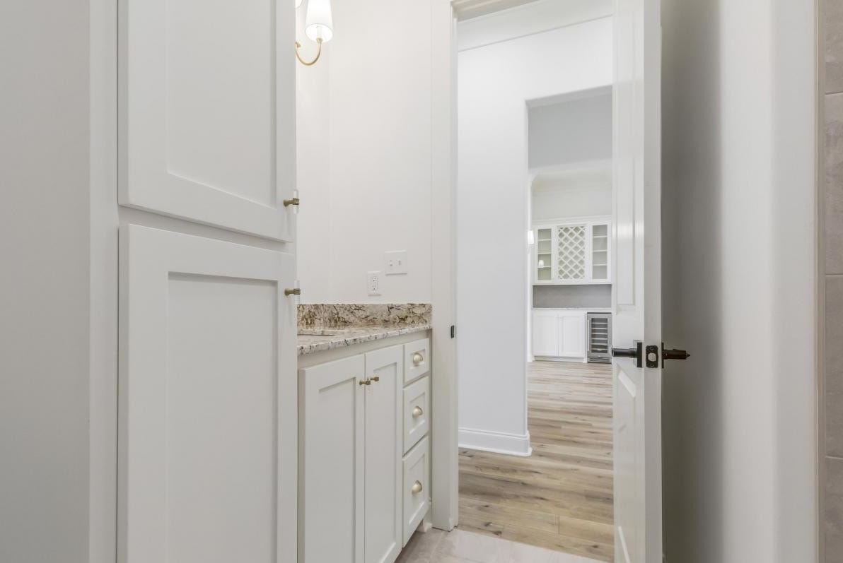 A small room with white cabinets, granite countertop, and light wood flooring, viewed through a partially open door. Another room with similar cabinetry and flooring is visible in the background.