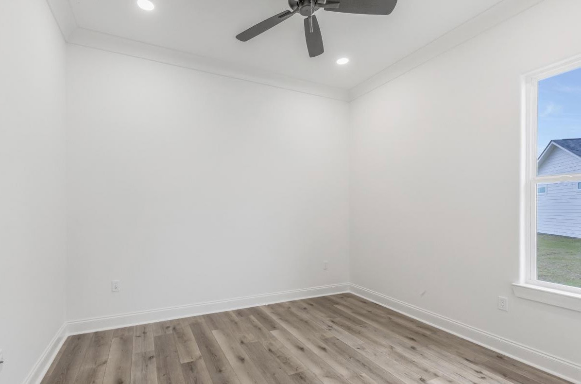 A small empty room with white walls, recessed ceiling lights, a ceiling fan, wood-look flooring, and a window showing part of a house and grass outside. The room is well-lit and unfurnished.