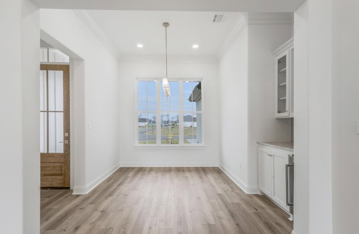 A bright, empty room with light wood flooring, white walls, a large window letting in natural light, a hanging light fixture, a wooden front door, and a built-in cabinet with open shelving on the right.
