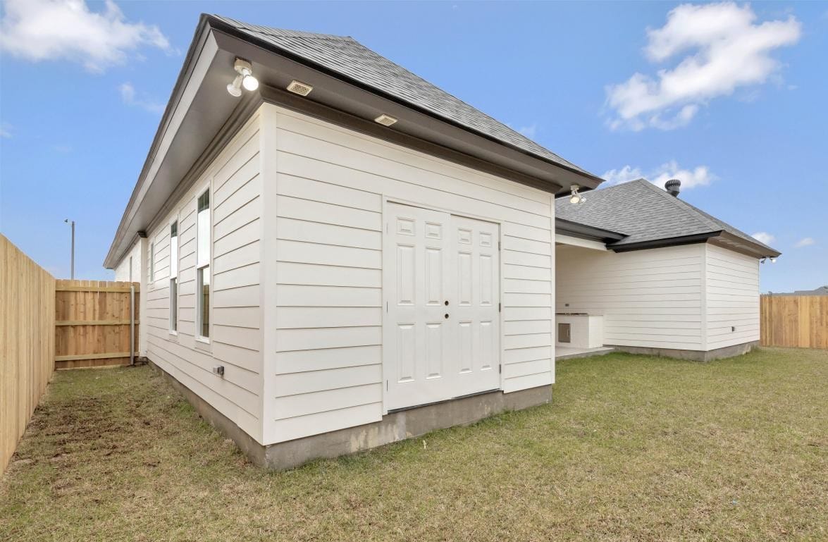 A white single-story house with horizontal siding and a gray shingled roof, featuring a double-door outdoor storage space and a fenced grass backyard under a blue sky with a few clouds.